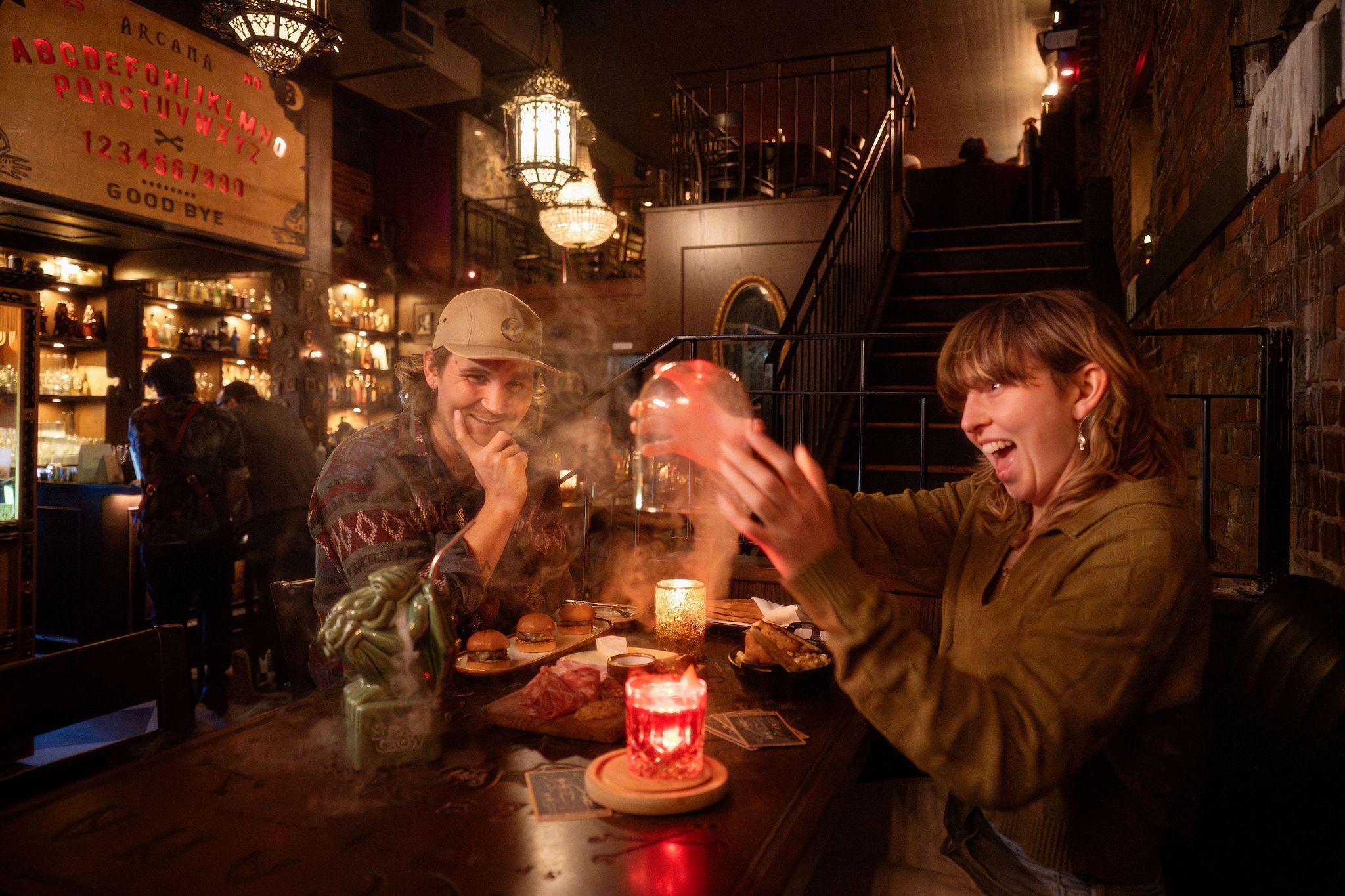 Two people at a dimly lit bar table at Arcana Food and Spirits with food and drinks, one person holding a glowing orb with smoke, the other smiling.