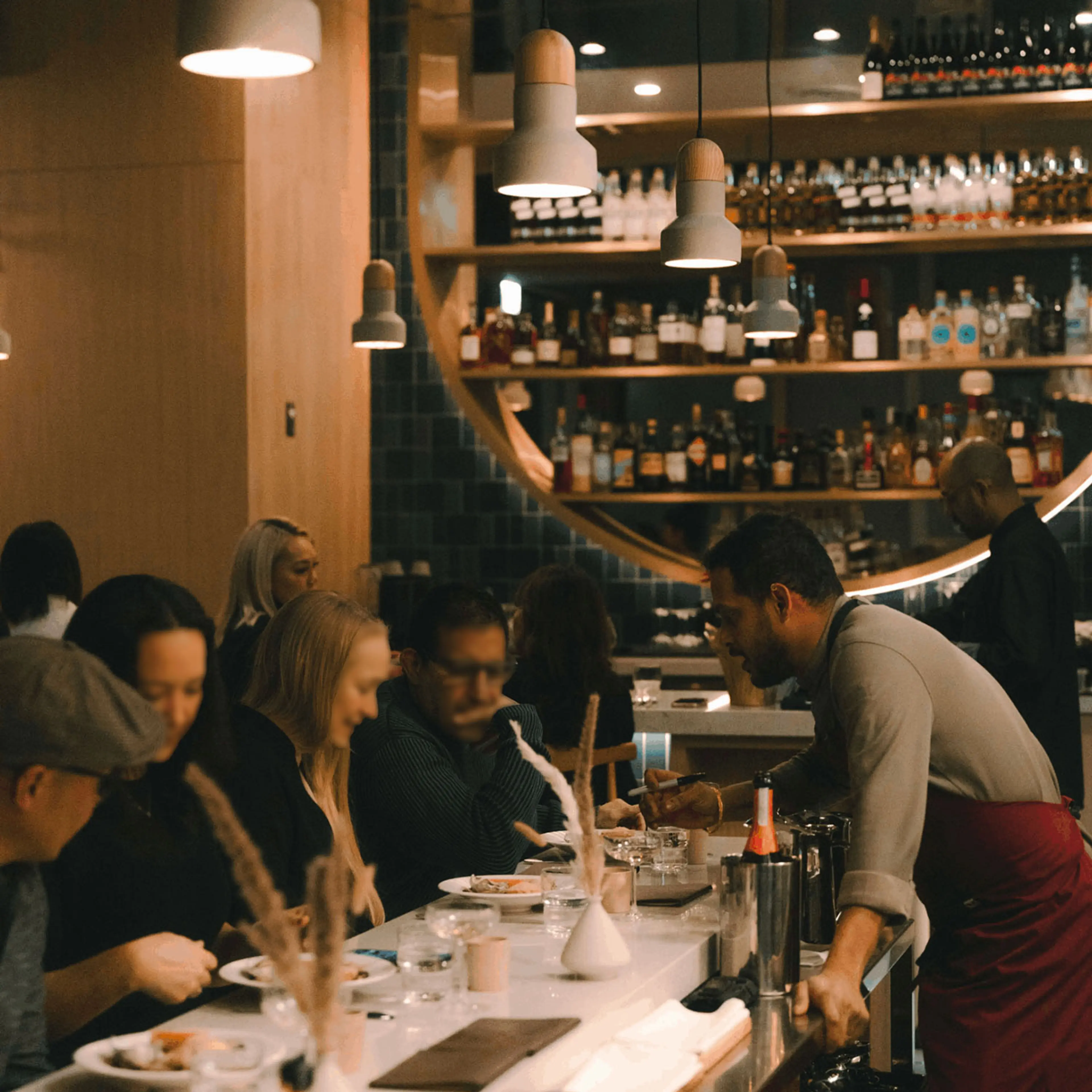 Bartender serving drinks to customers seated at Kavita's bar counter with shelves of bottles in the background.