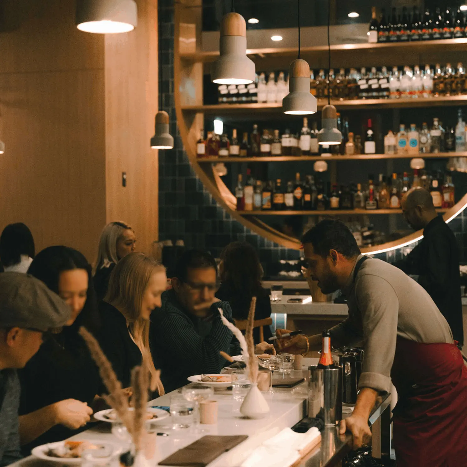 Bartender serving drinks to customers seated at Kavita's bar counter with shelves of bottles in the background.