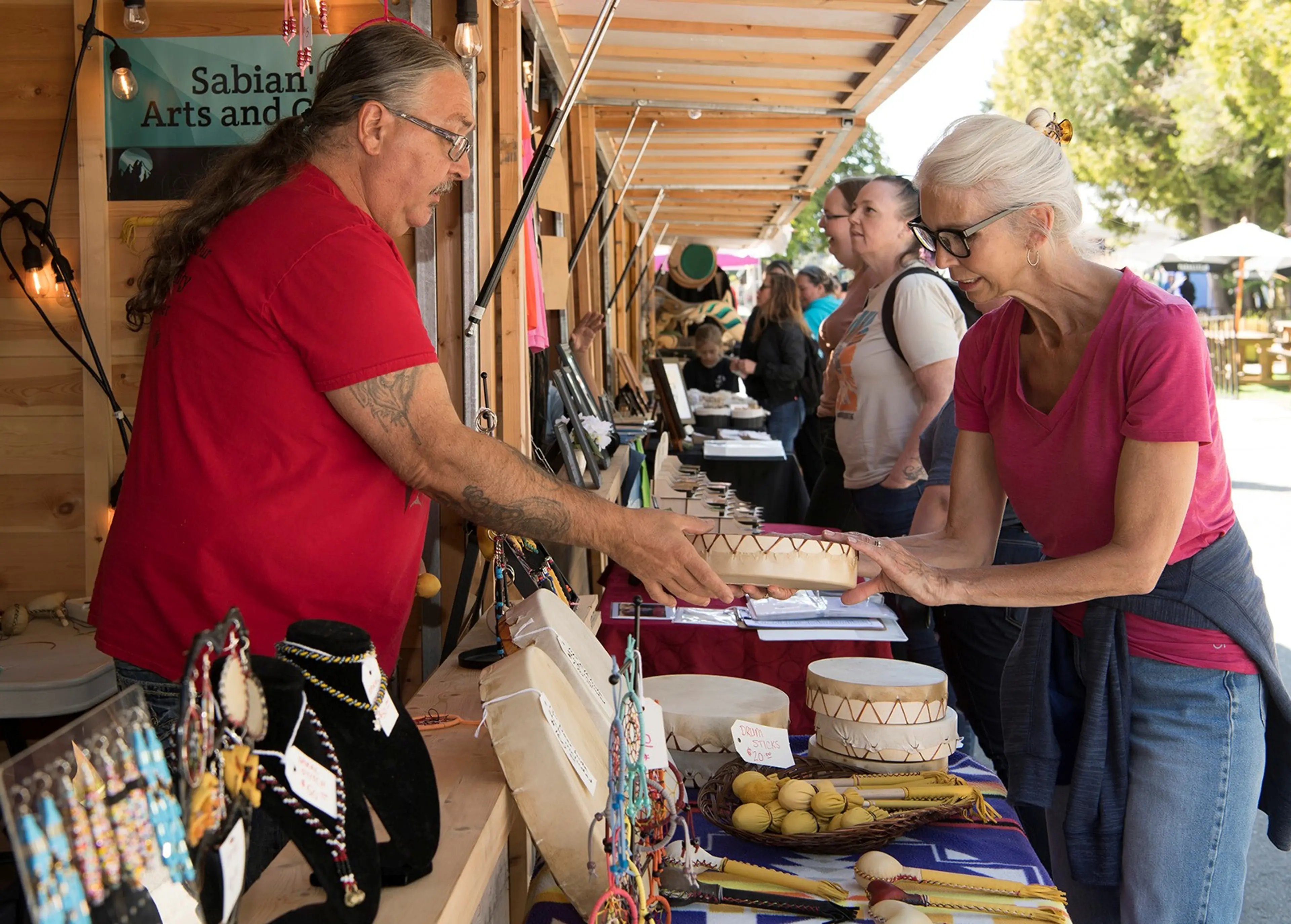 A person in a red shirt hands a drum to a person in a pink shirt at an outdoor market stall with jewelry and drums displayed.