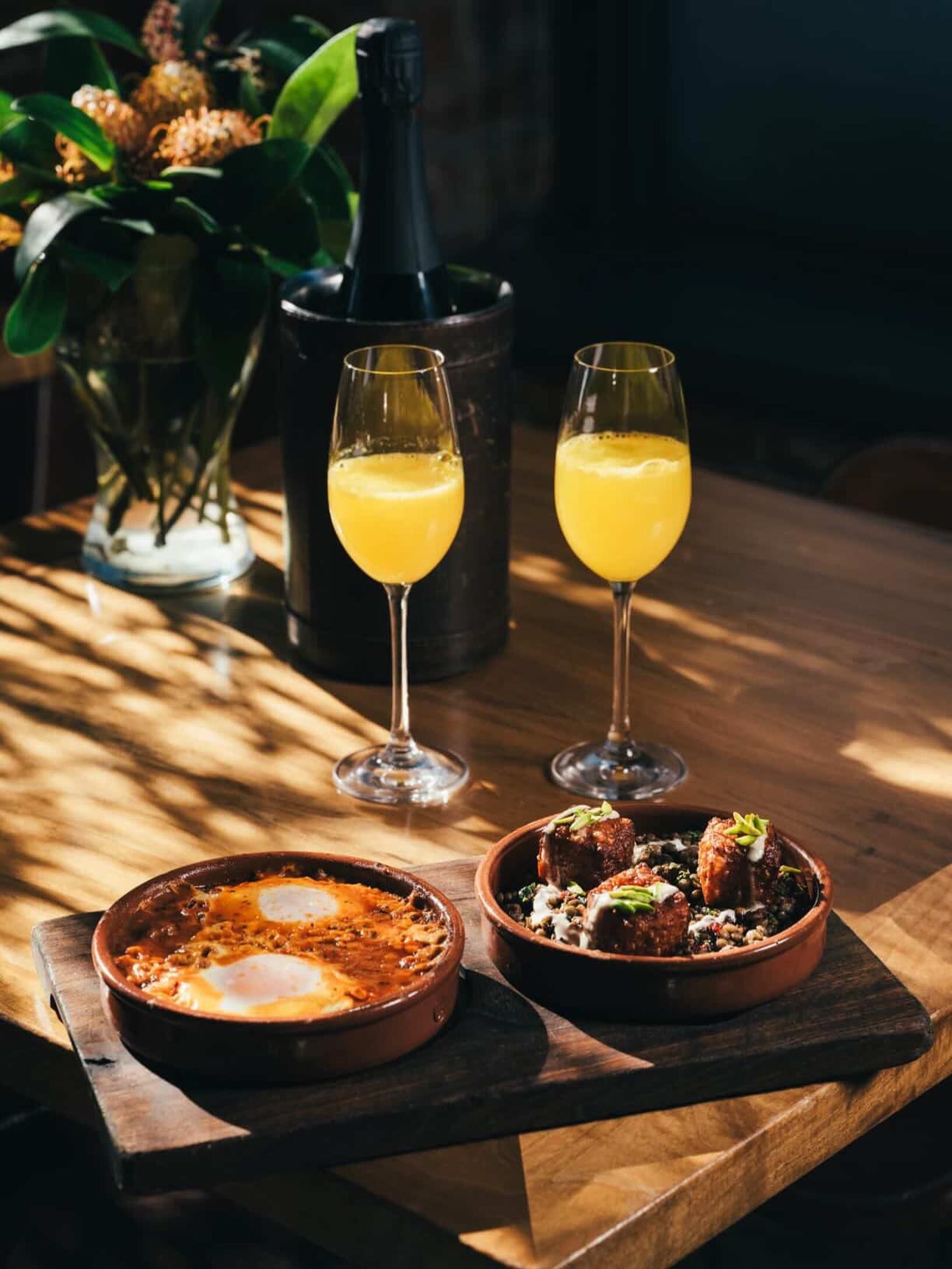 Two glasses of orange juice, a bottle in an ice bucket, and two dishes with eggs and garnished food on a wooden table.