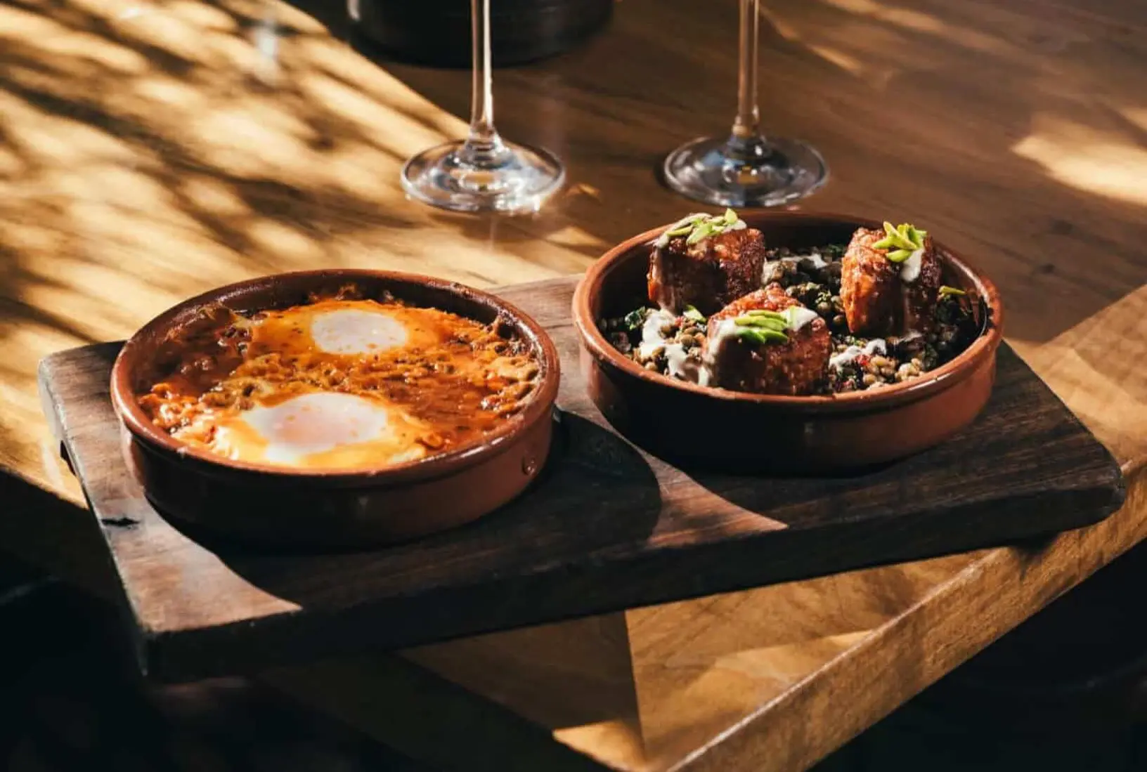 Two glasses of orange juice, a bottle in an ice bucket, and two dishes with eggs and garnished food on a wooden table.