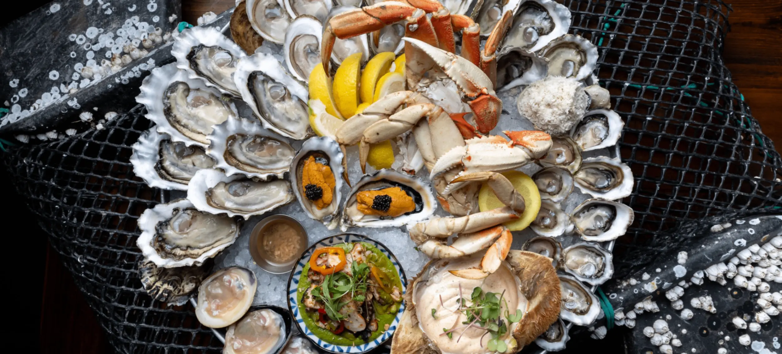 Seafood platter with crab legs, oysters, clams, lemon wedges, and sauces on a bed of ice over a fishing net.