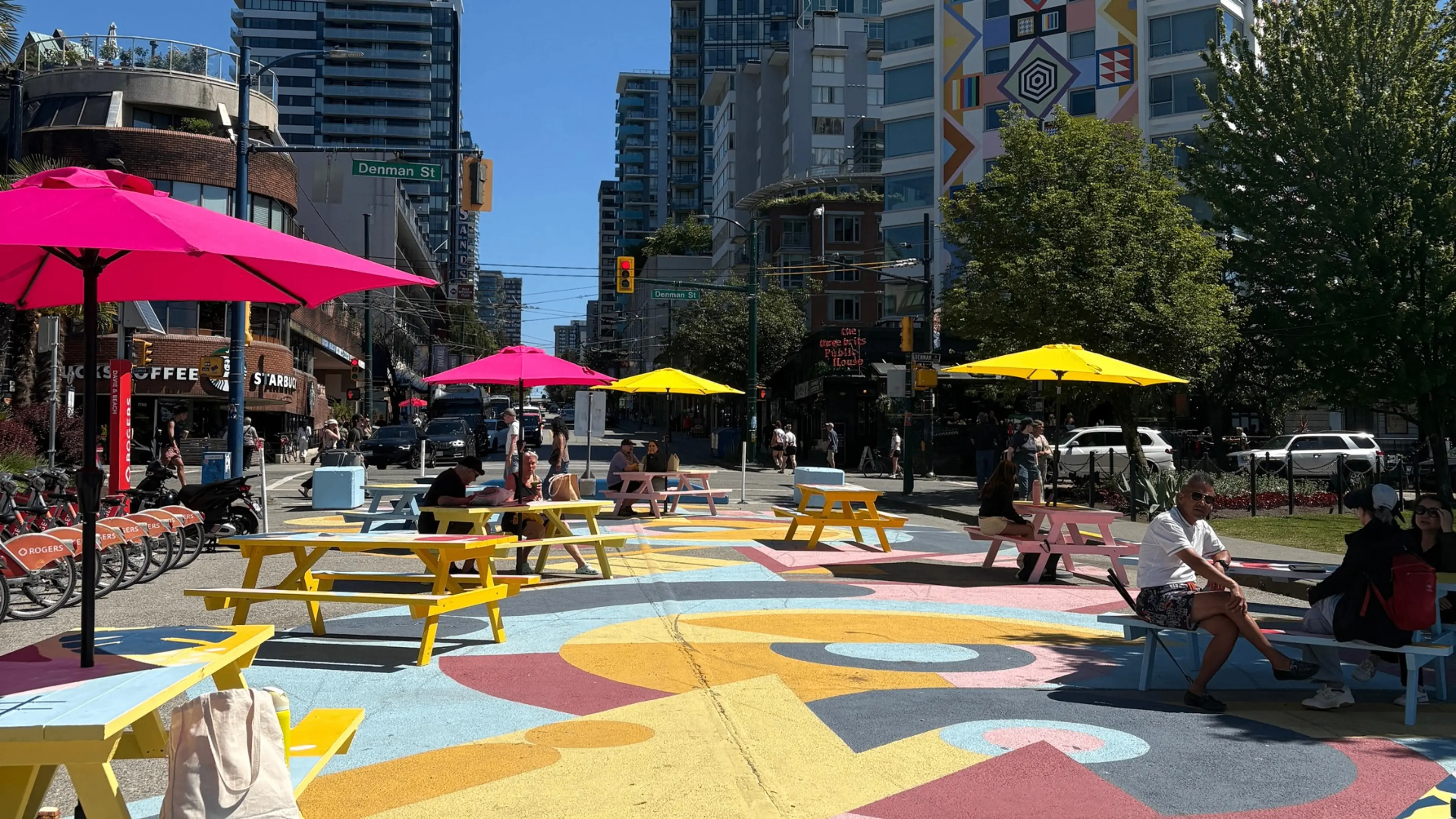 Outdoor urban seating area with colorful picnic tables and umbrellas on a painted street with people sitting and walking.