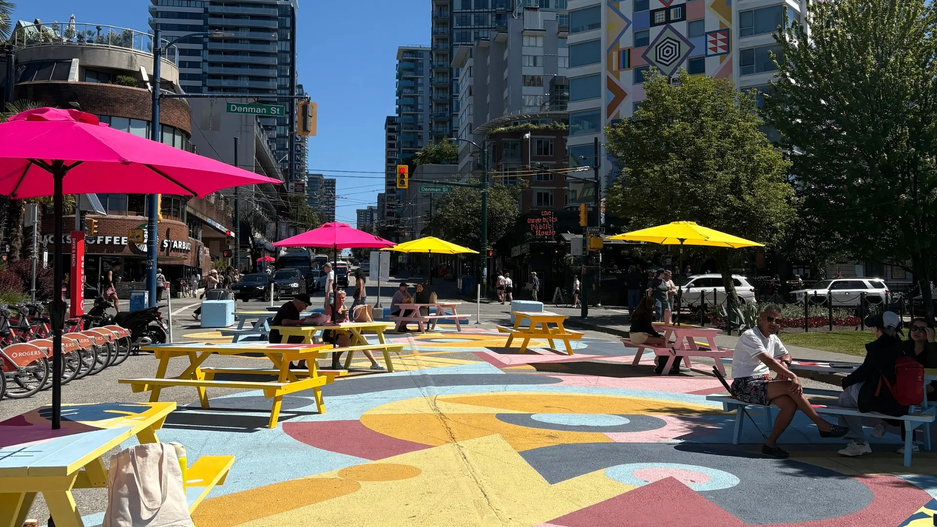 Outdoor urban seating area with colorful picnic tables and umbrellas on a painted street with people sitting and walking.