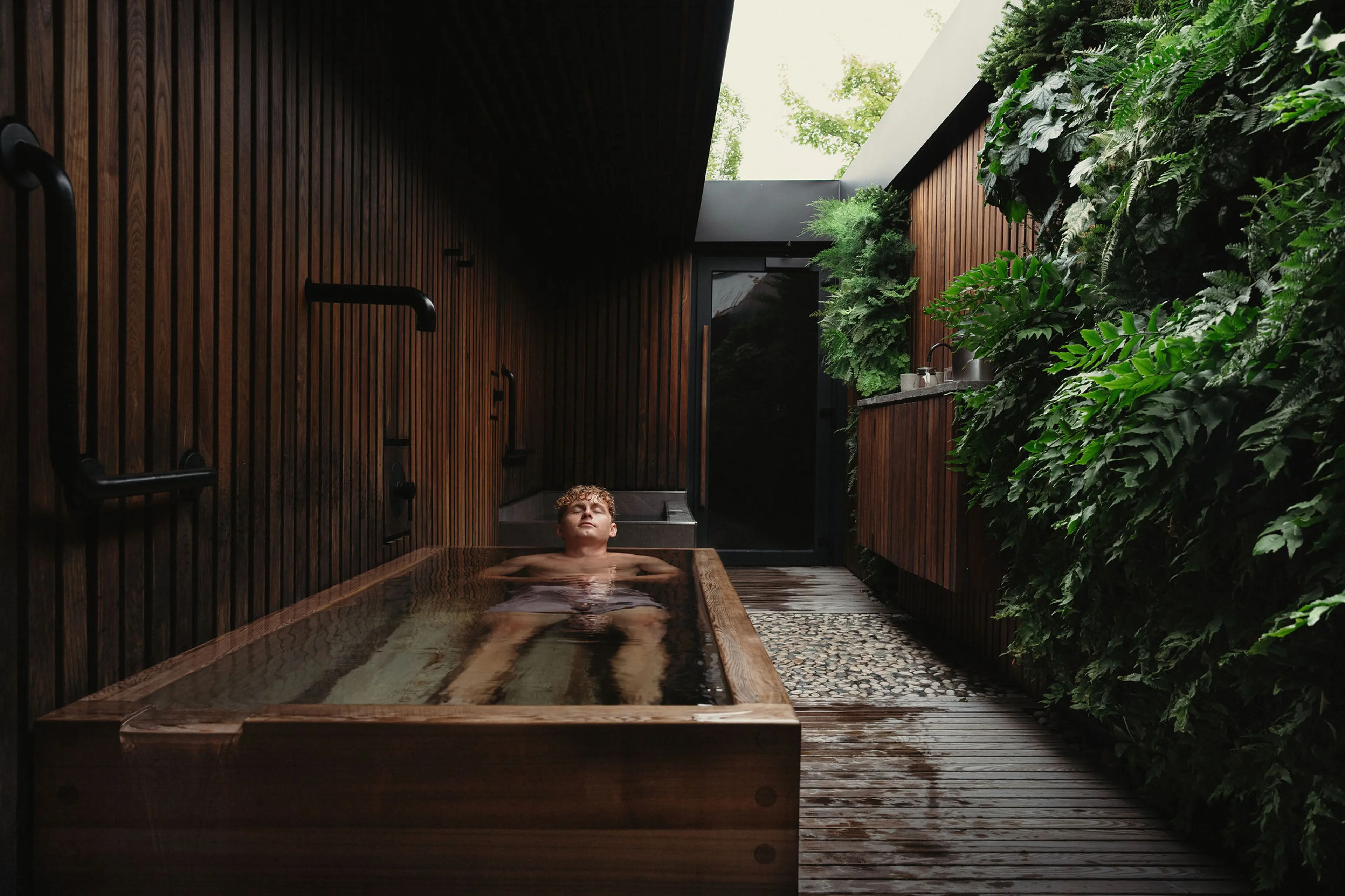 Person relaxing in a wooden bathtub with water flowing, next to a green vertical garden wall.
