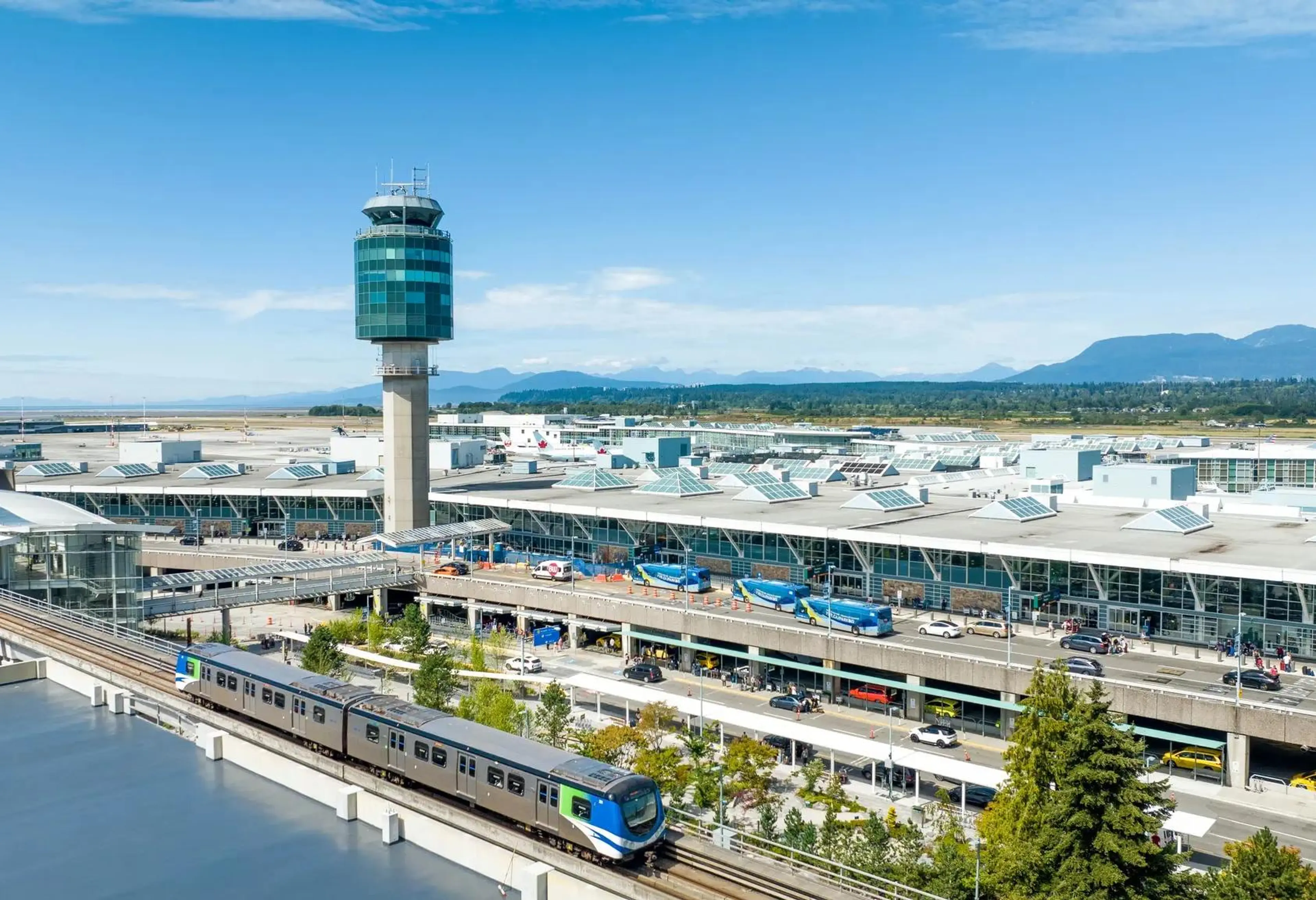 YVR Airport terminal with control tower, buses, cars, and a train on elevated tracks under a clear blue sky.