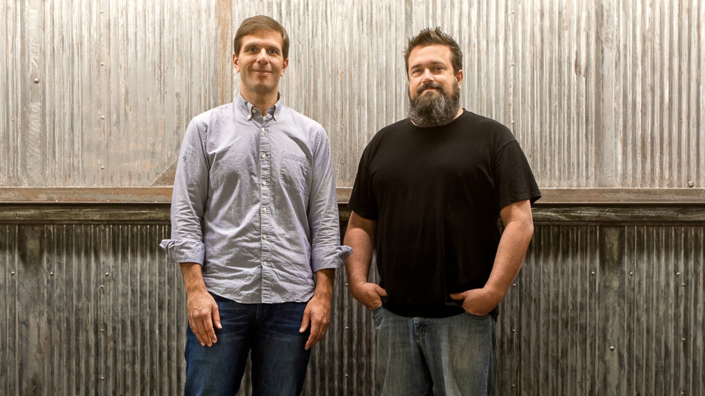 Two people standing side by side against a corrugated metal wall, one wearing a light button-up shirt and jeans, the other in a black t-shirt and jeans.