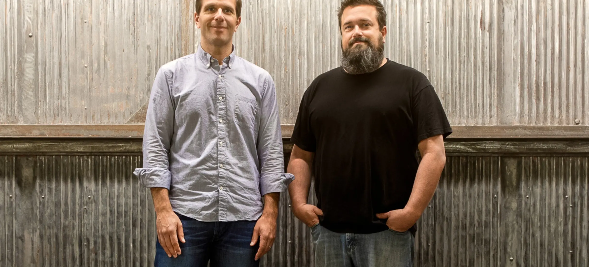 Two people standing side by side against a corrugated metal wall, one wearing a light button-up shirt and jeans, the other in a black t-shirt and jeans.