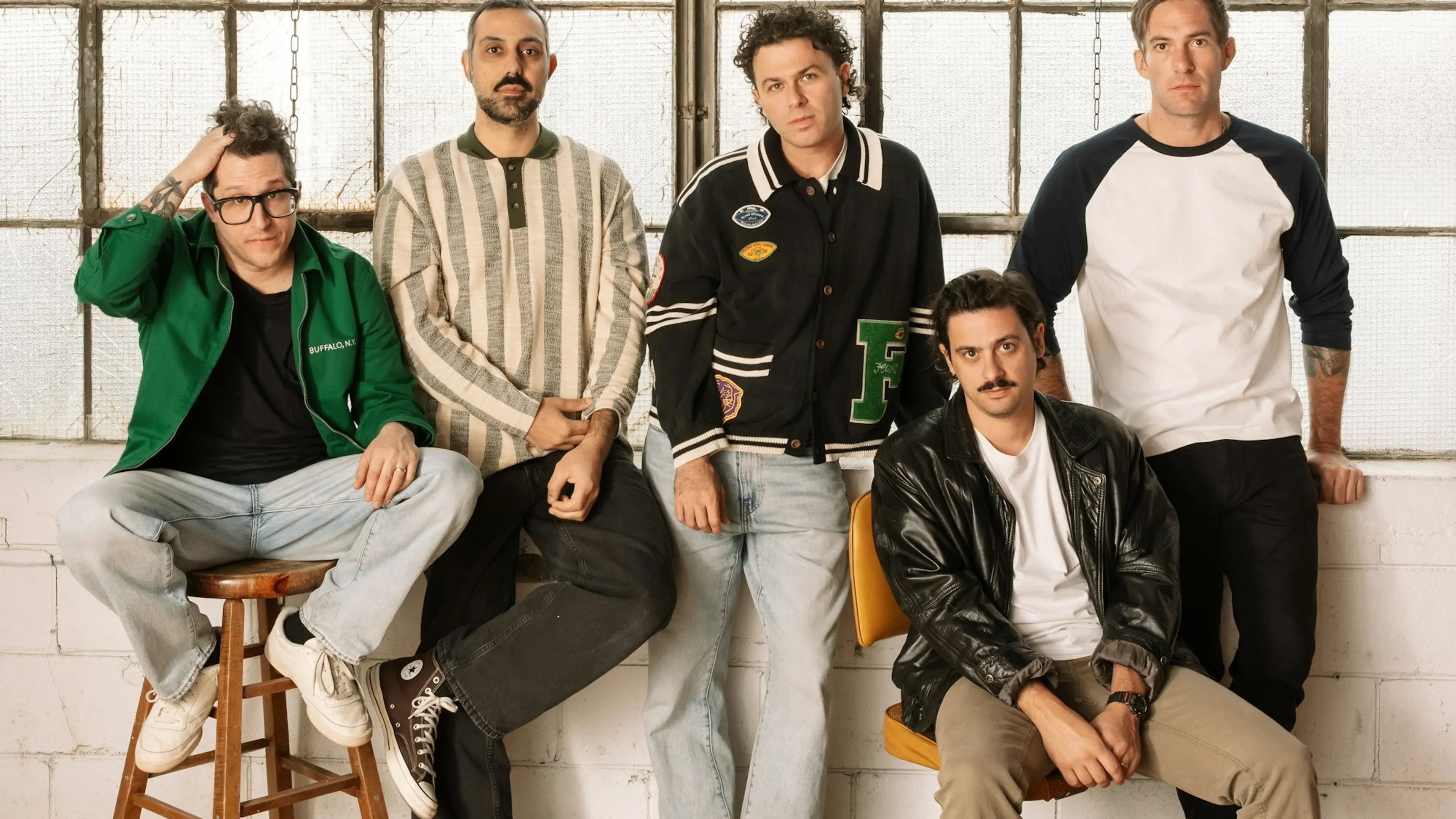 Five people posing indoors against a window, three standing and two sitting on stools, wearing casual clothes including jackets and sweaters.