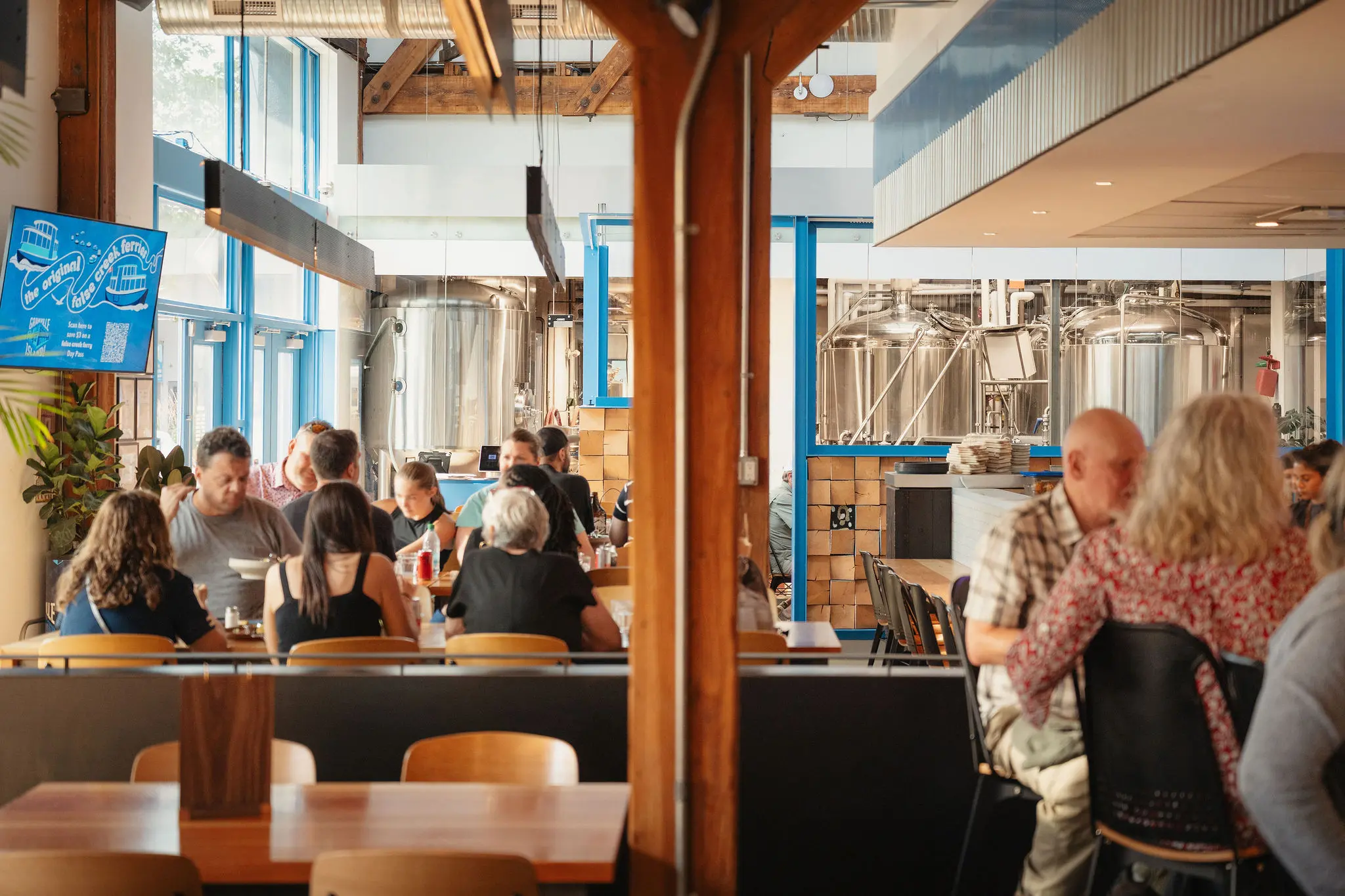 People sitting and dining in a bright brewery restaurant with large metal brewing tanks visible through windows.