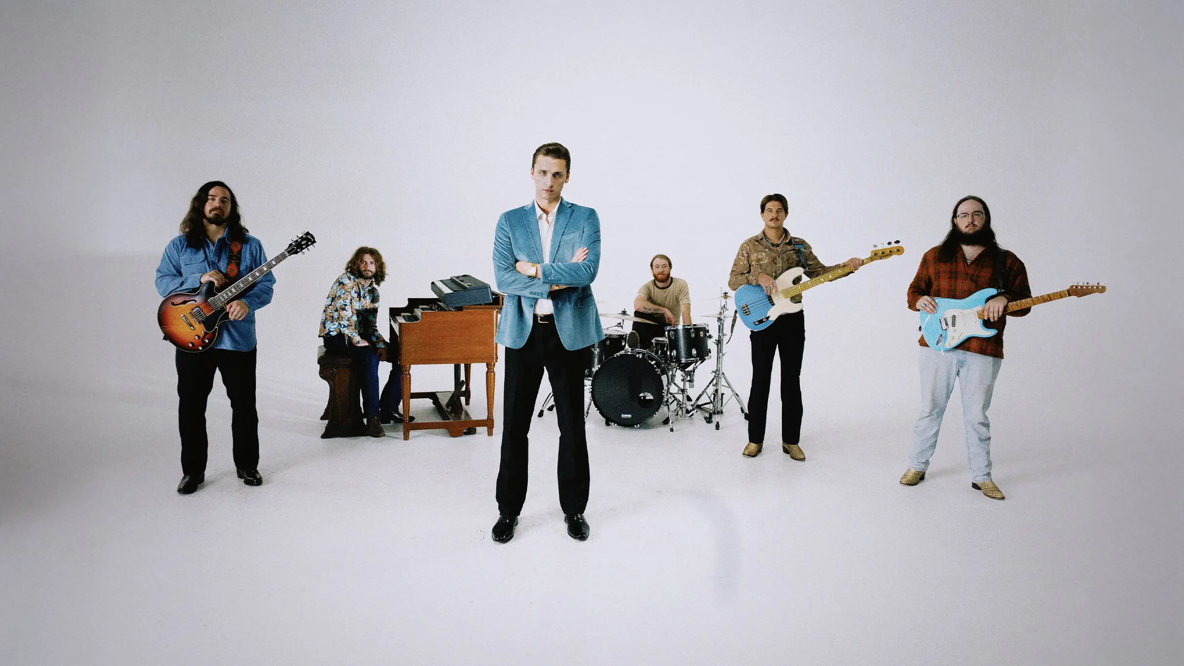Six people in a studio with musical instruments including guitars, keyboard, and drums, standing and sitting against a white background.