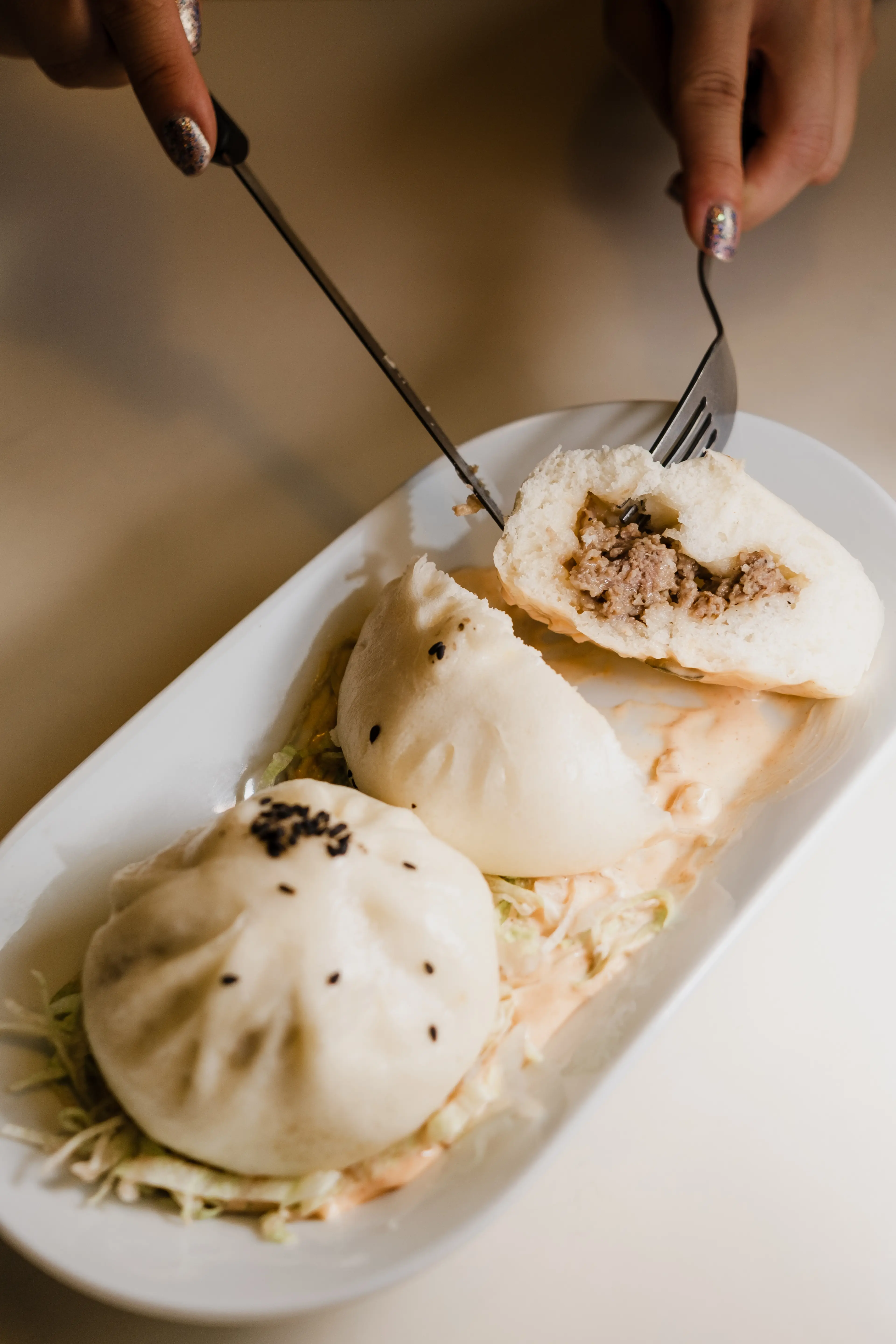 Two hands using a fork and knife to cut a steamed bun filled with minced meat on a white plate with two other buns.
