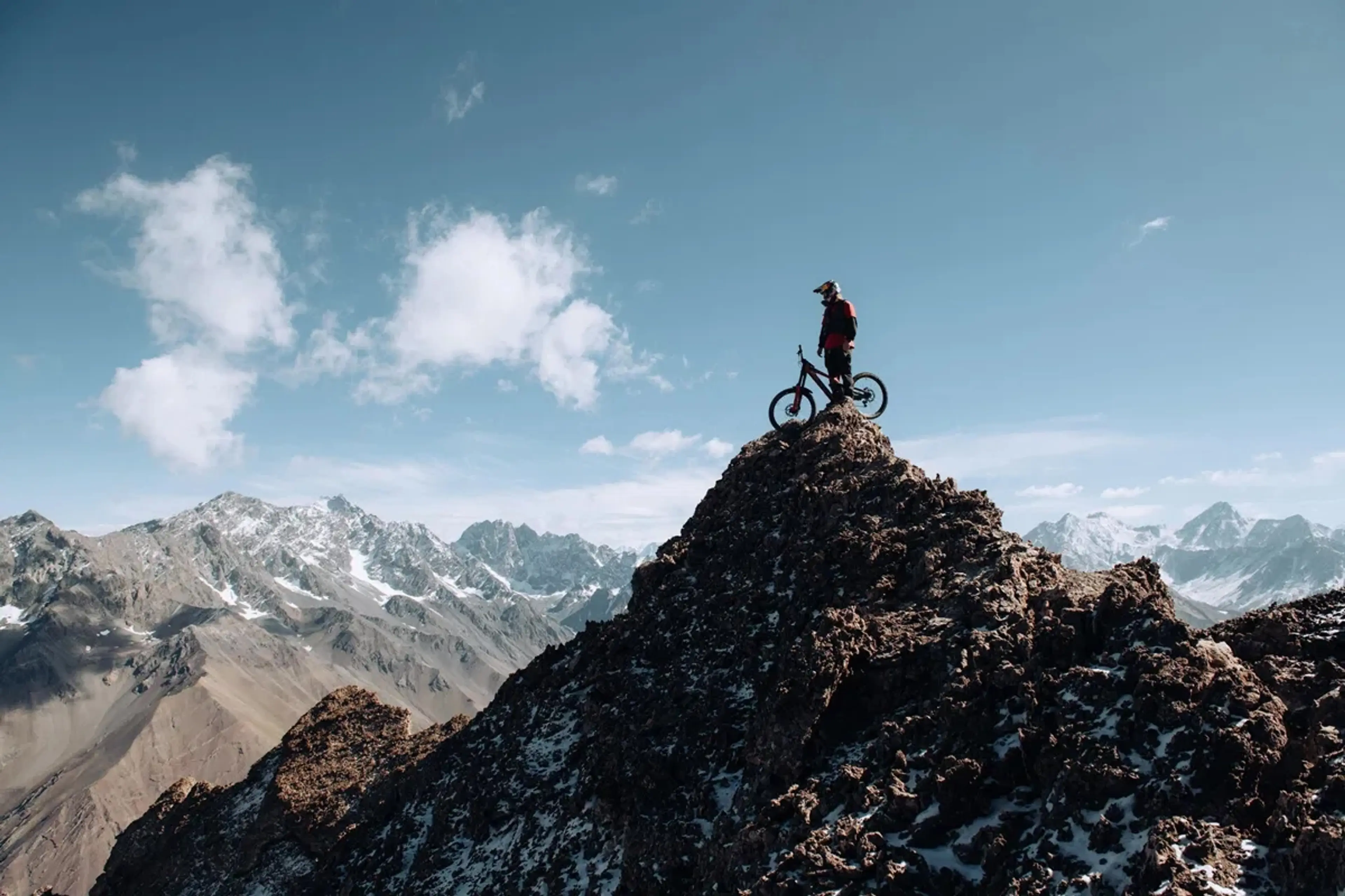A man stands next to mountain bike on top of a rocky peak with a dusting of snow in a still from the film Anytime.