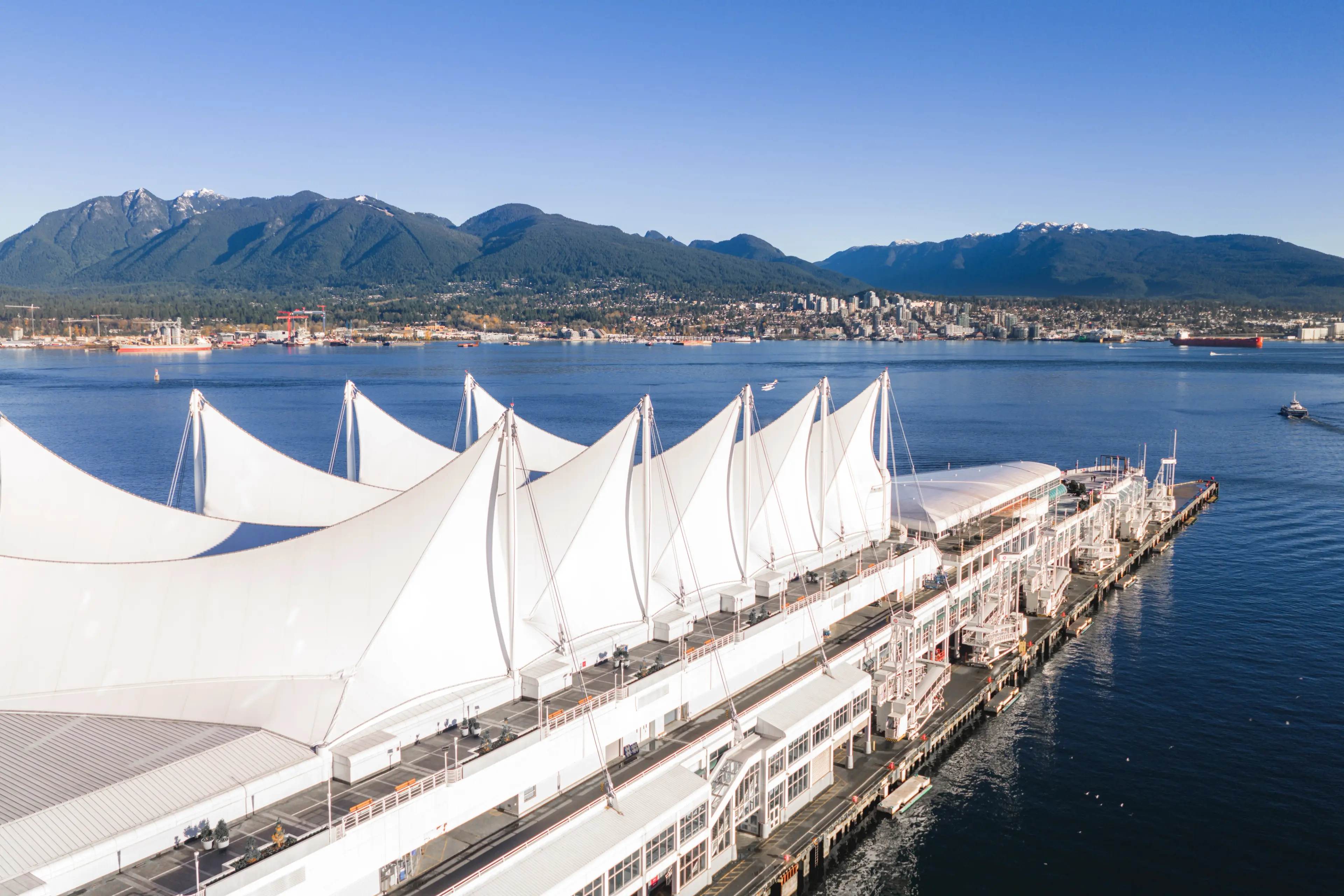 Waterfront building with white sail-like roof structures, mountains and city skyline in the background under clear blue sky.