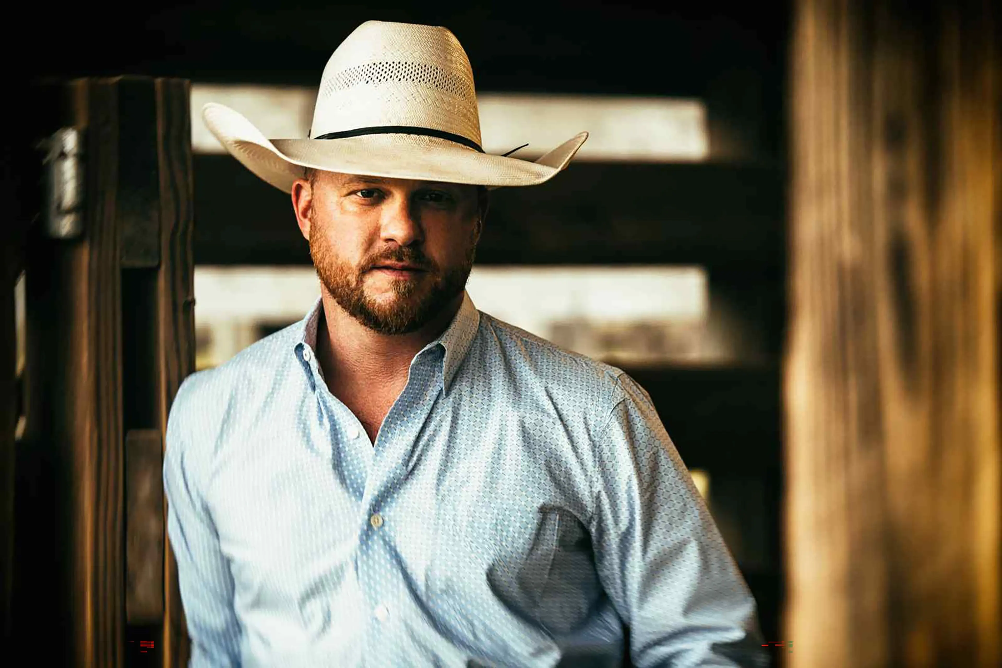 Person wearing a white cowboy hat and light blue button-up shirt standing near wooden beams in a dimly lit setting.