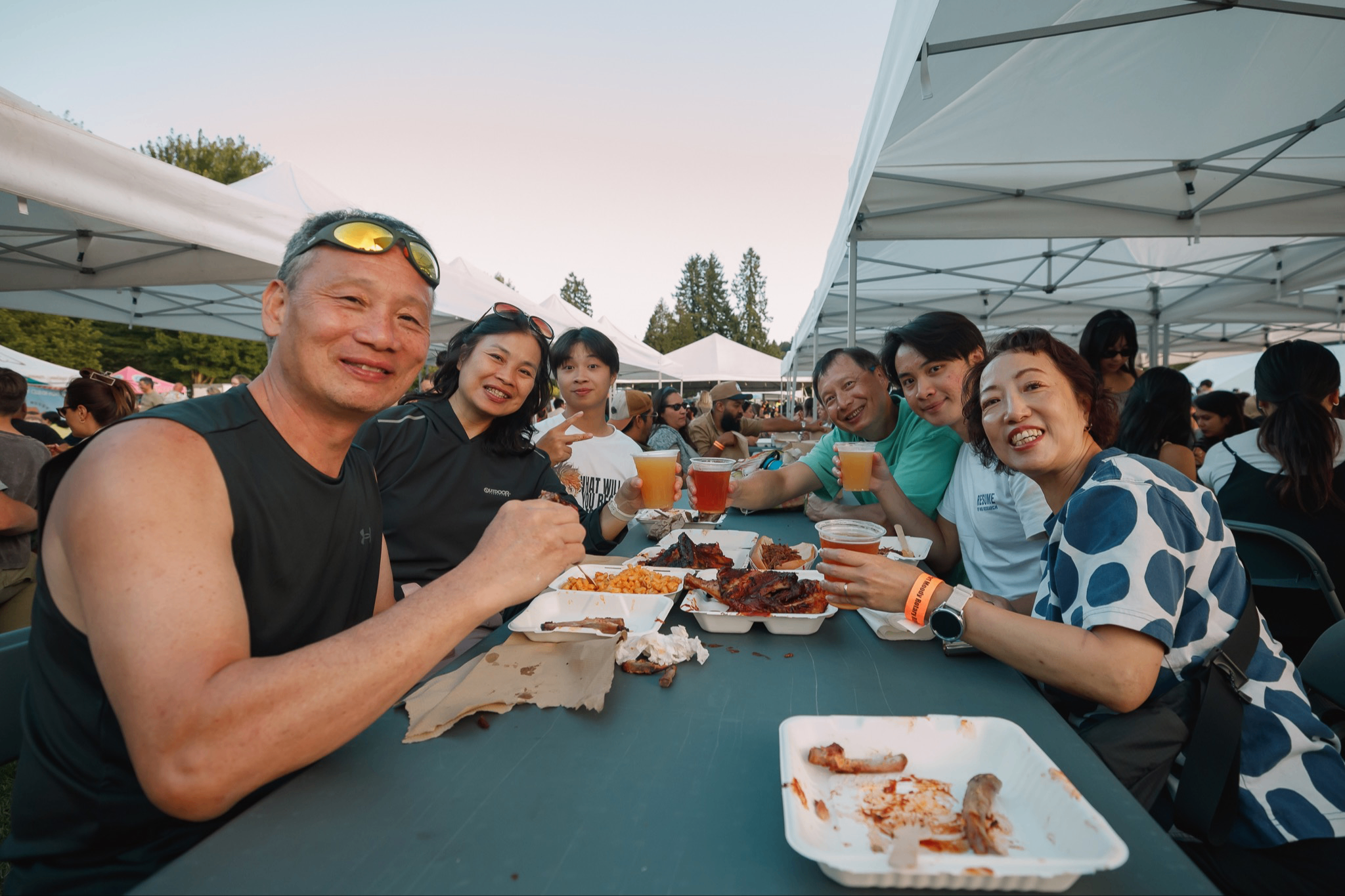 Six people sitting at a picnic table under white tents, eating food and holding drinks, smiling at the camera.