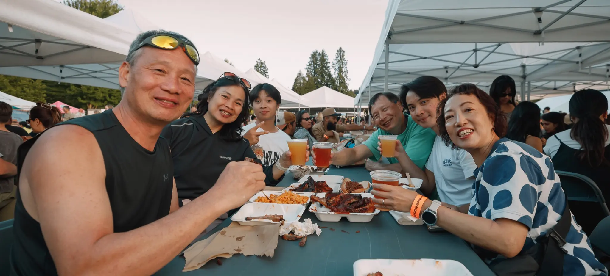 Six people sitting at a picnic table under white tents, eating food and holding drinks, smiling at the camera.