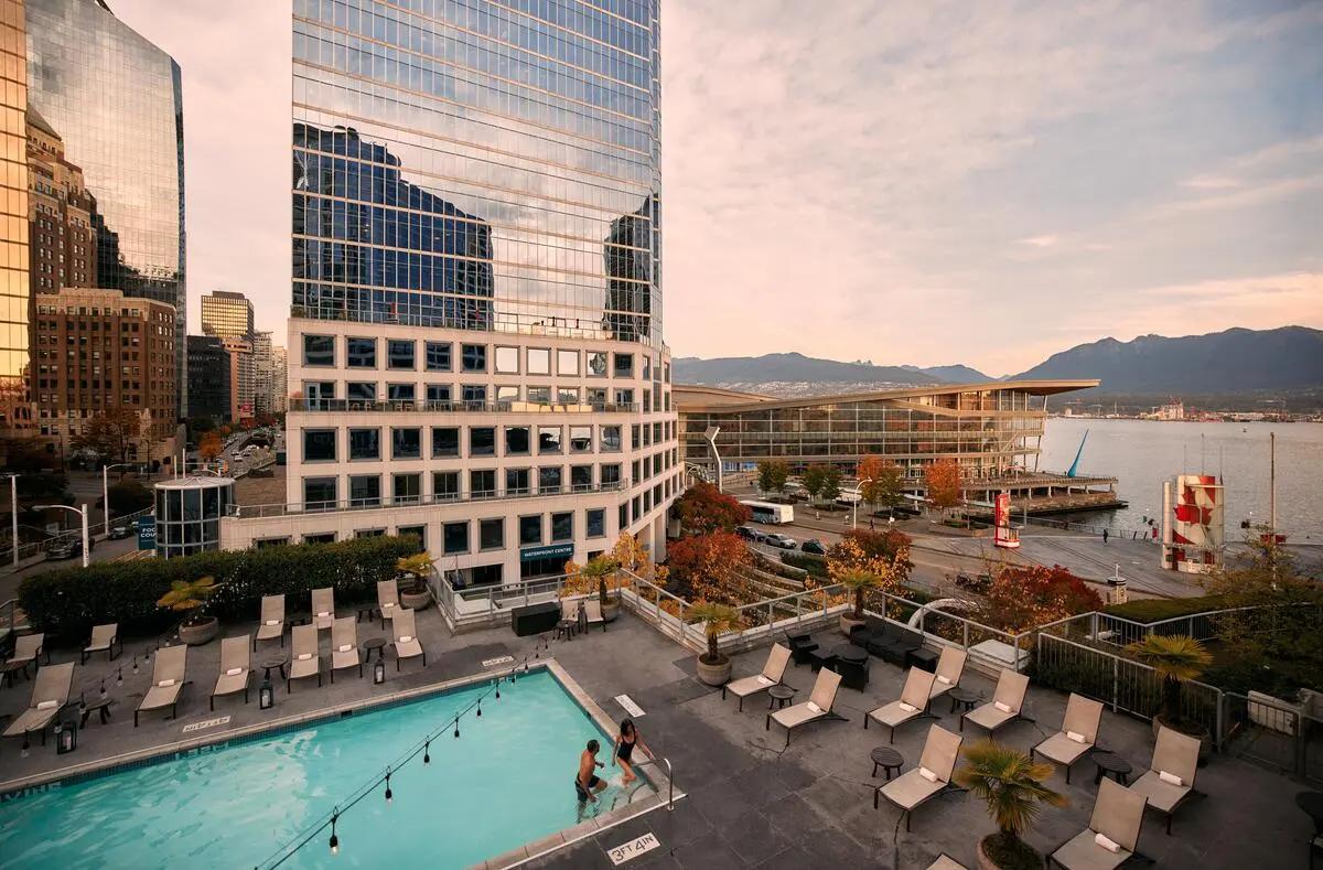 Outdoor pool with two people entering, surrounded by lounge chairs, near tall glass buildings and waterfront with mountains in the background.