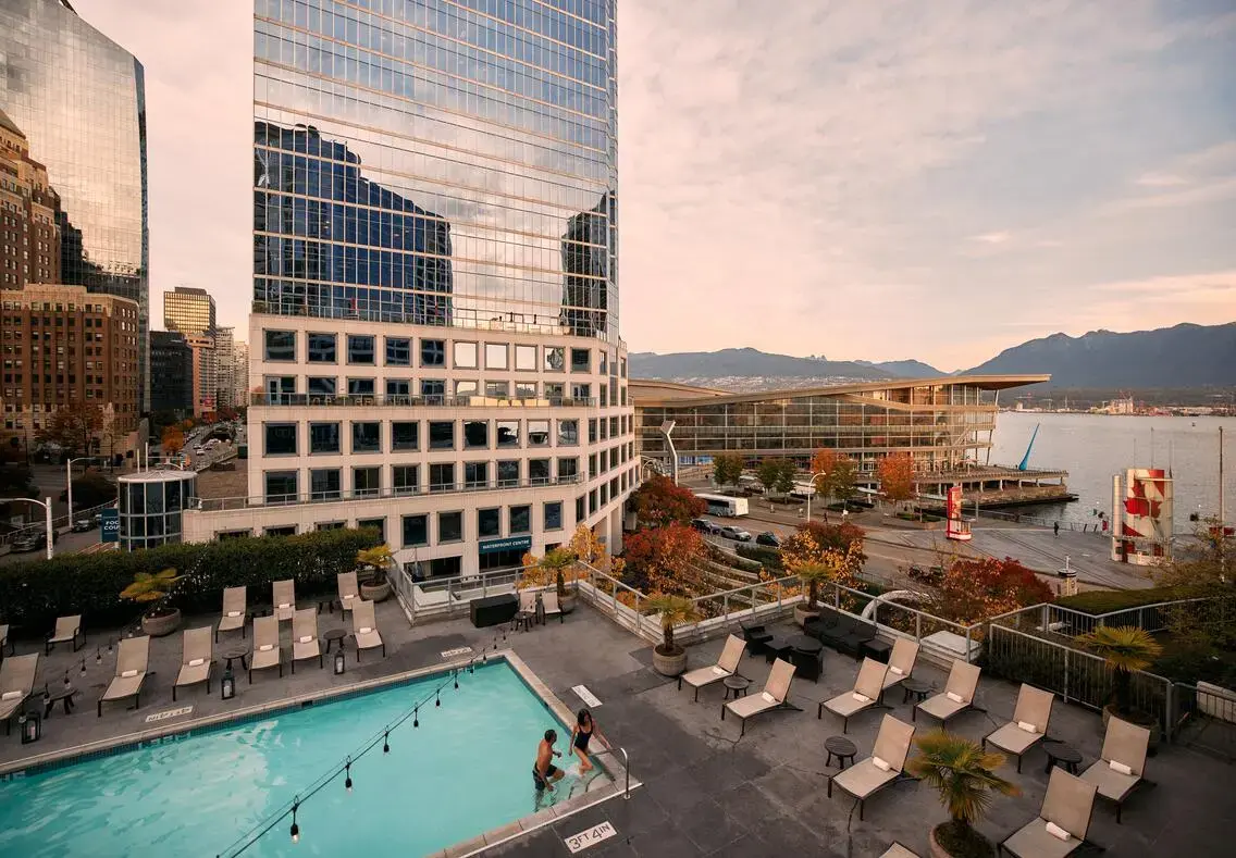 Outdoor pool with two people entering, surrounded by lounge chairs, near tall glass buildings and waterfront with mountains in the background.