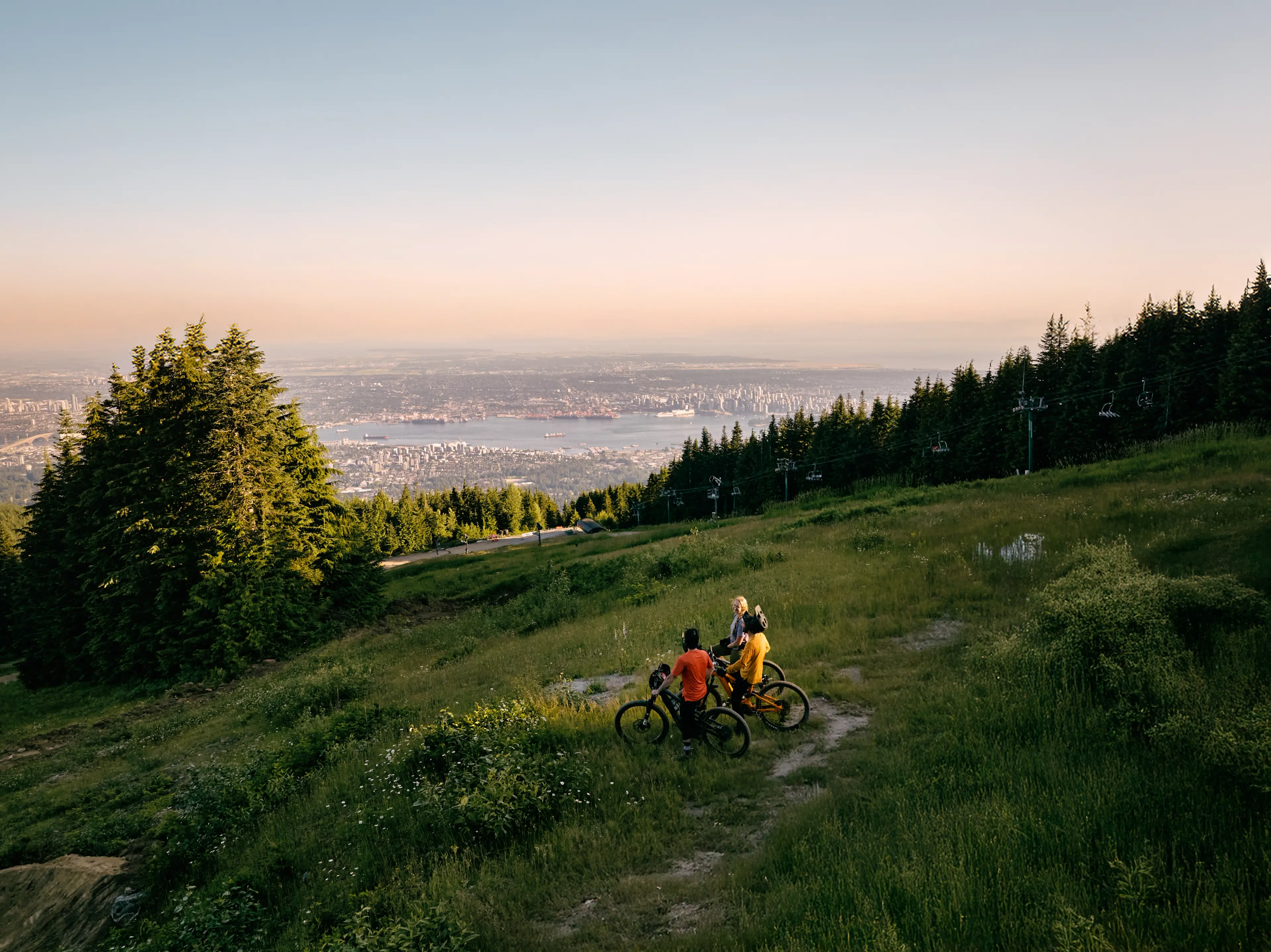 Two people with bicycles on a grassy hillside at Grouse Mountain overlooking Vancouver and the ocean at sunset.