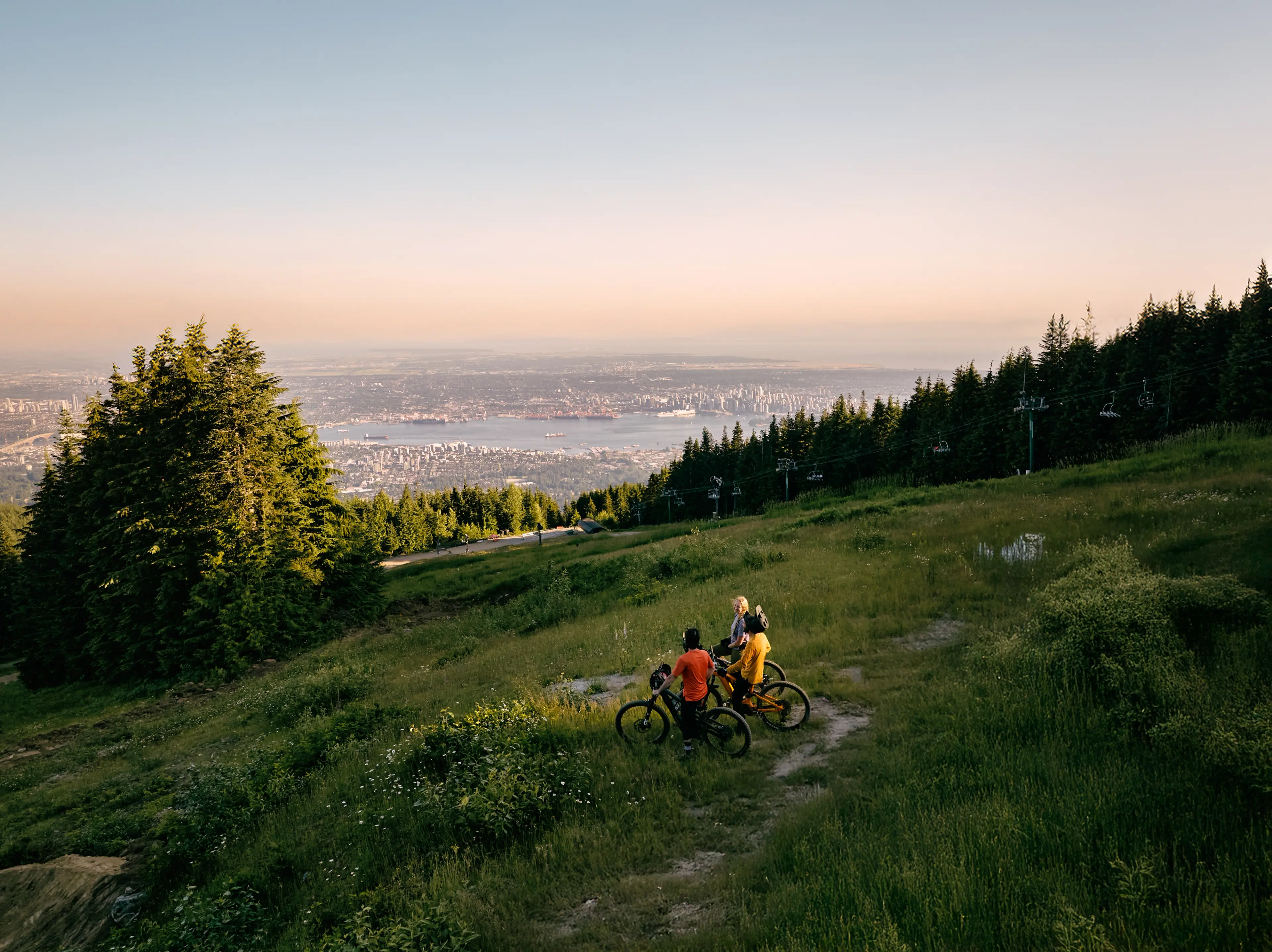 Two people with bicycles on a grassy hillside at Grouse Mountain overlooking Vancouver and the ocean at sunset.