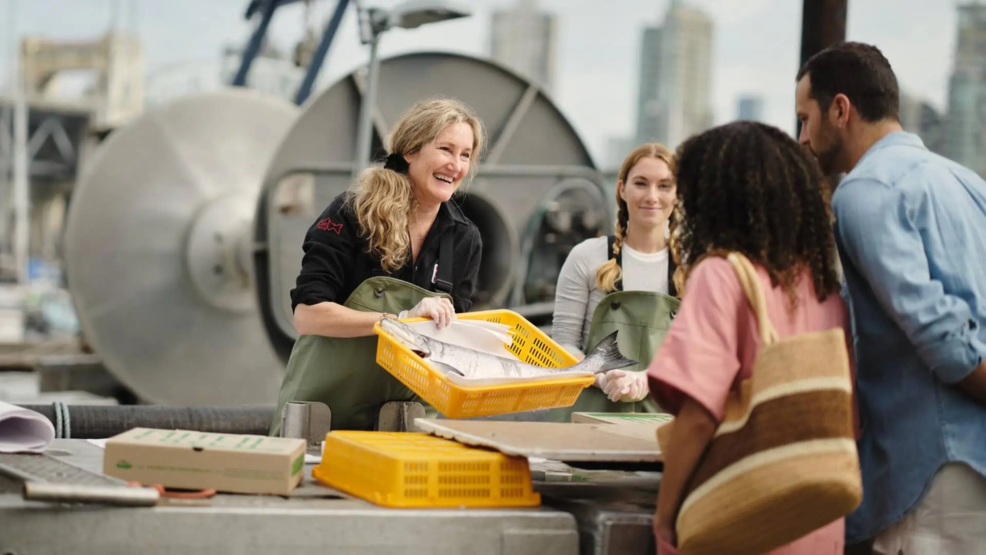 Two women in green aprons holding a basket of fish, smiling at a man and woman standing across a table outdoors.