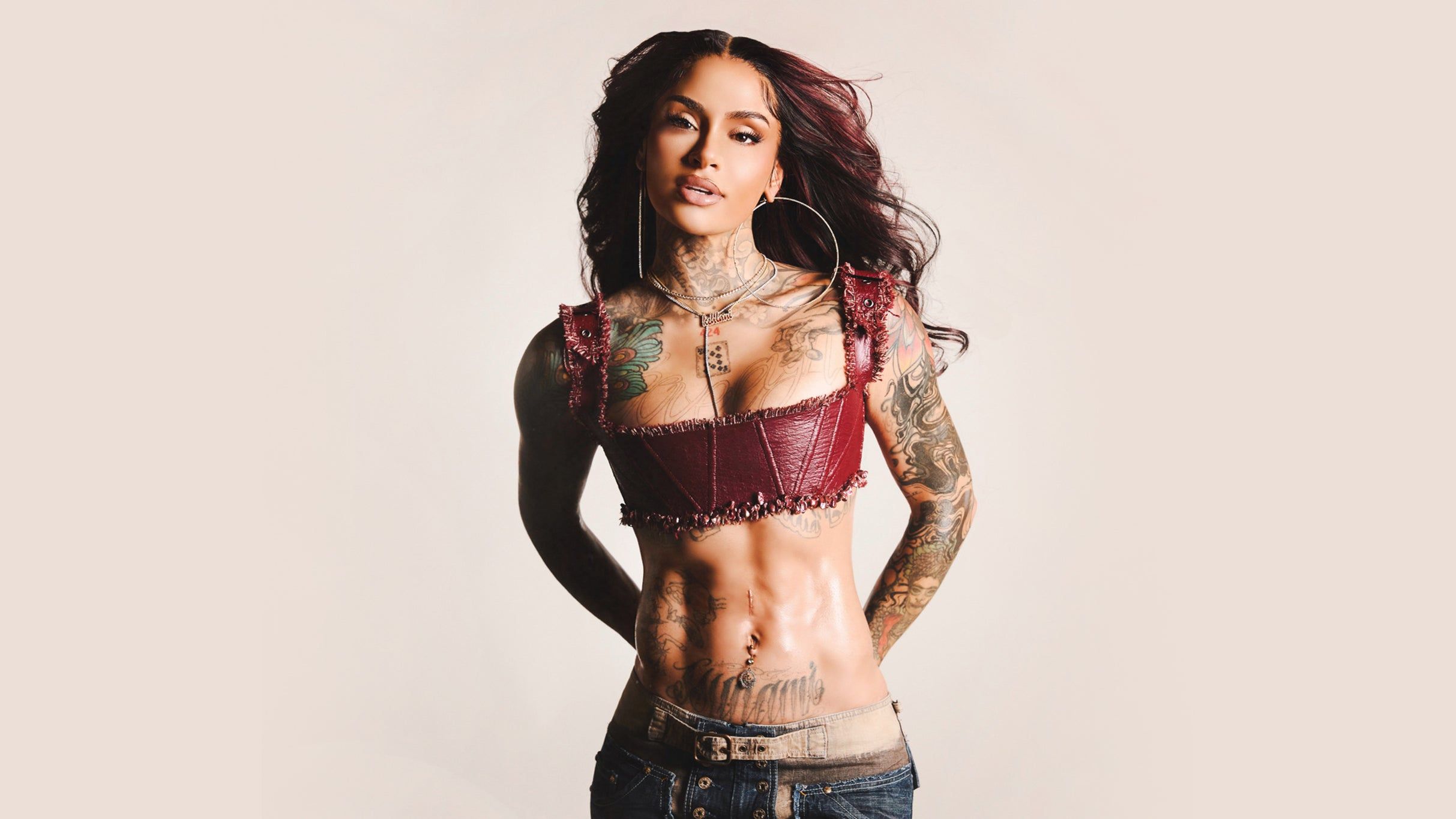 Person with long dark hair wearing a maroon crop top and jeans, showing tattoos on arms, chest, and abdomen.