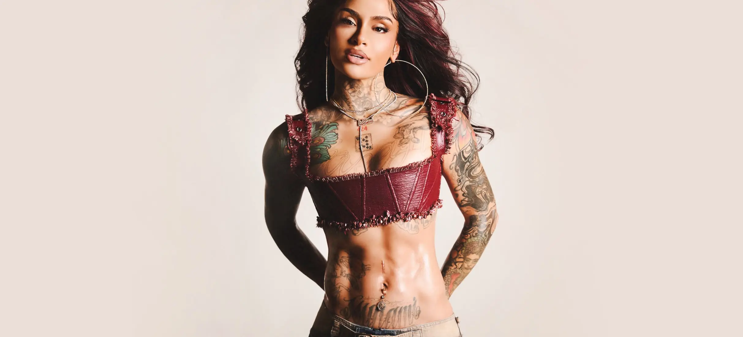 Person with long dark hair wearing a maroon crop top and jeans, showing tattoos on arms, chest, and abdomen.