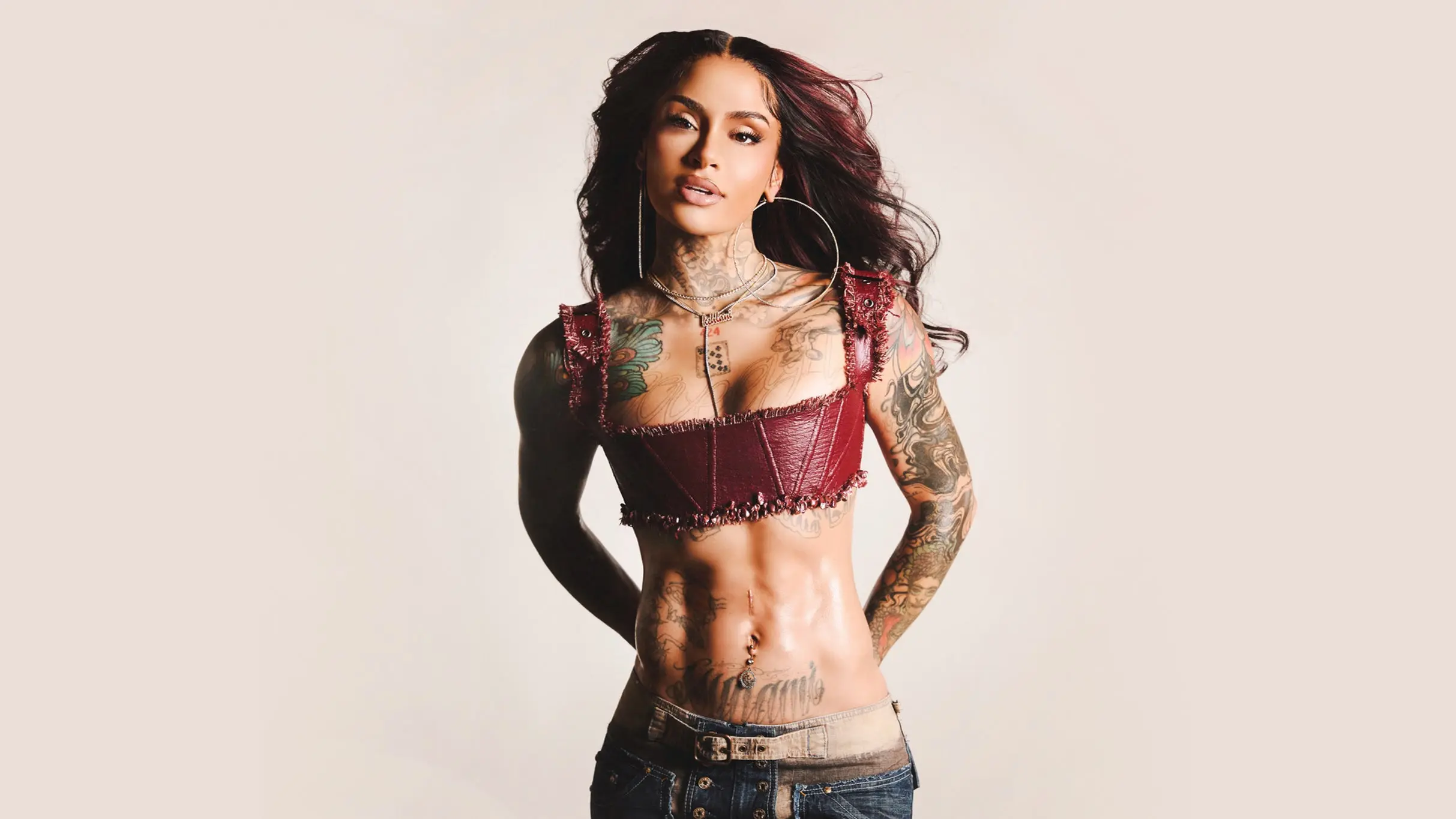 Person with long dark hair wearing a maroon crop top and jeans, showing tattoos on arms, chest, and abdomen.