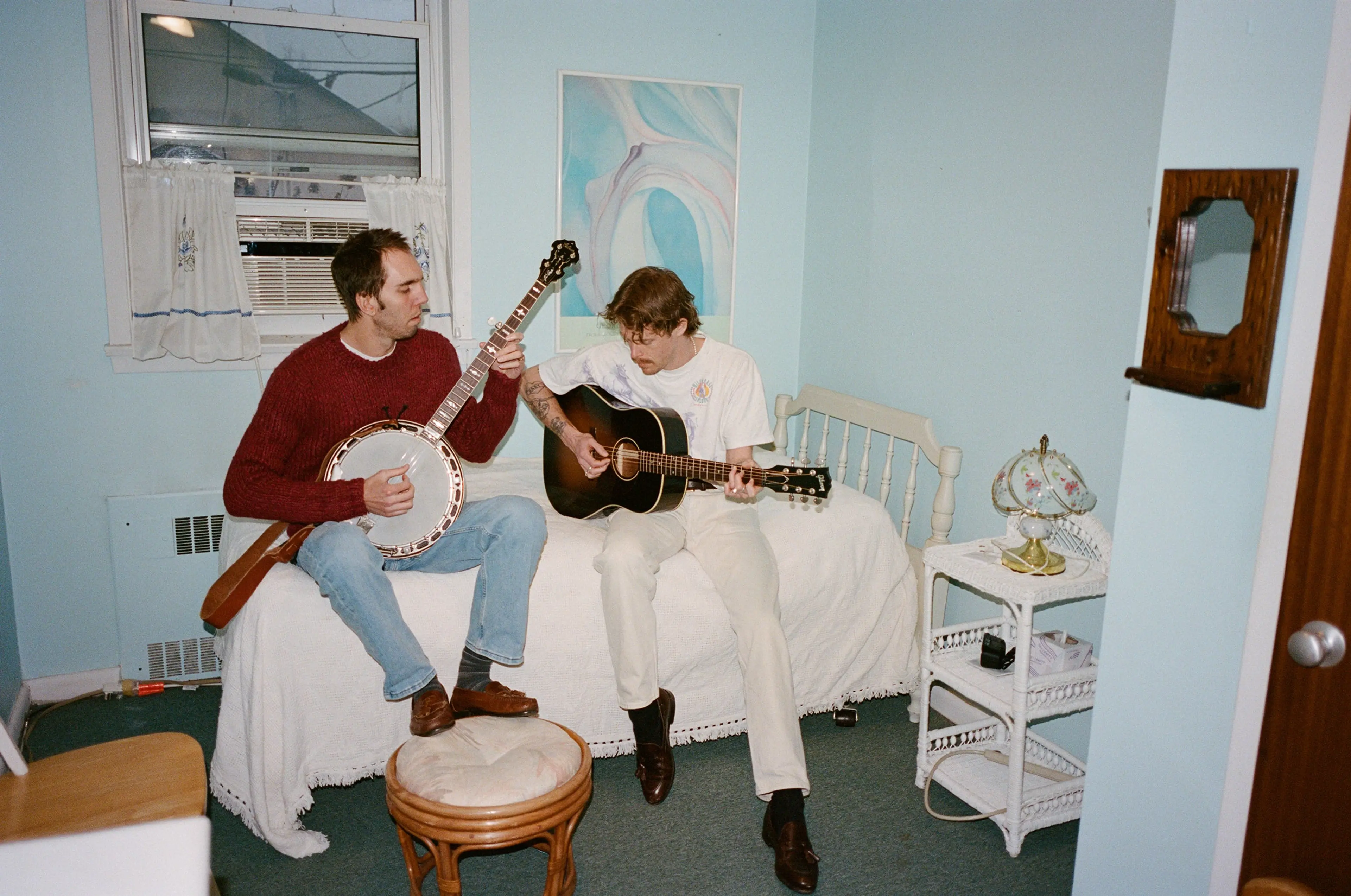 Two people sitting on a bed in a blue room playing banjo and acoustic guitar.