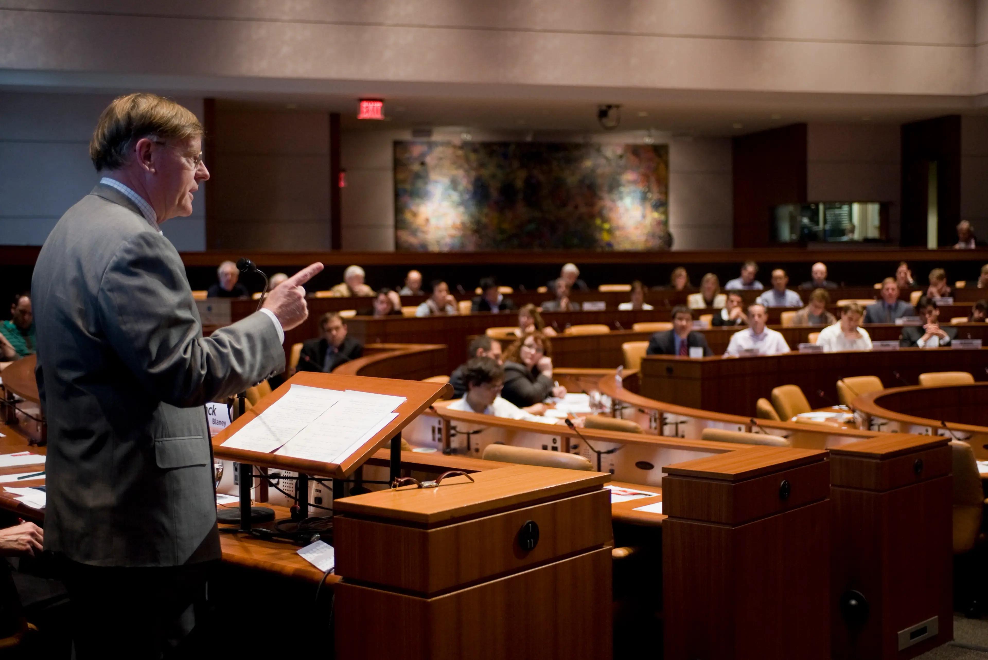 A person in a suit speaking at a podium in a large conference room with an audience seated in curved rows.