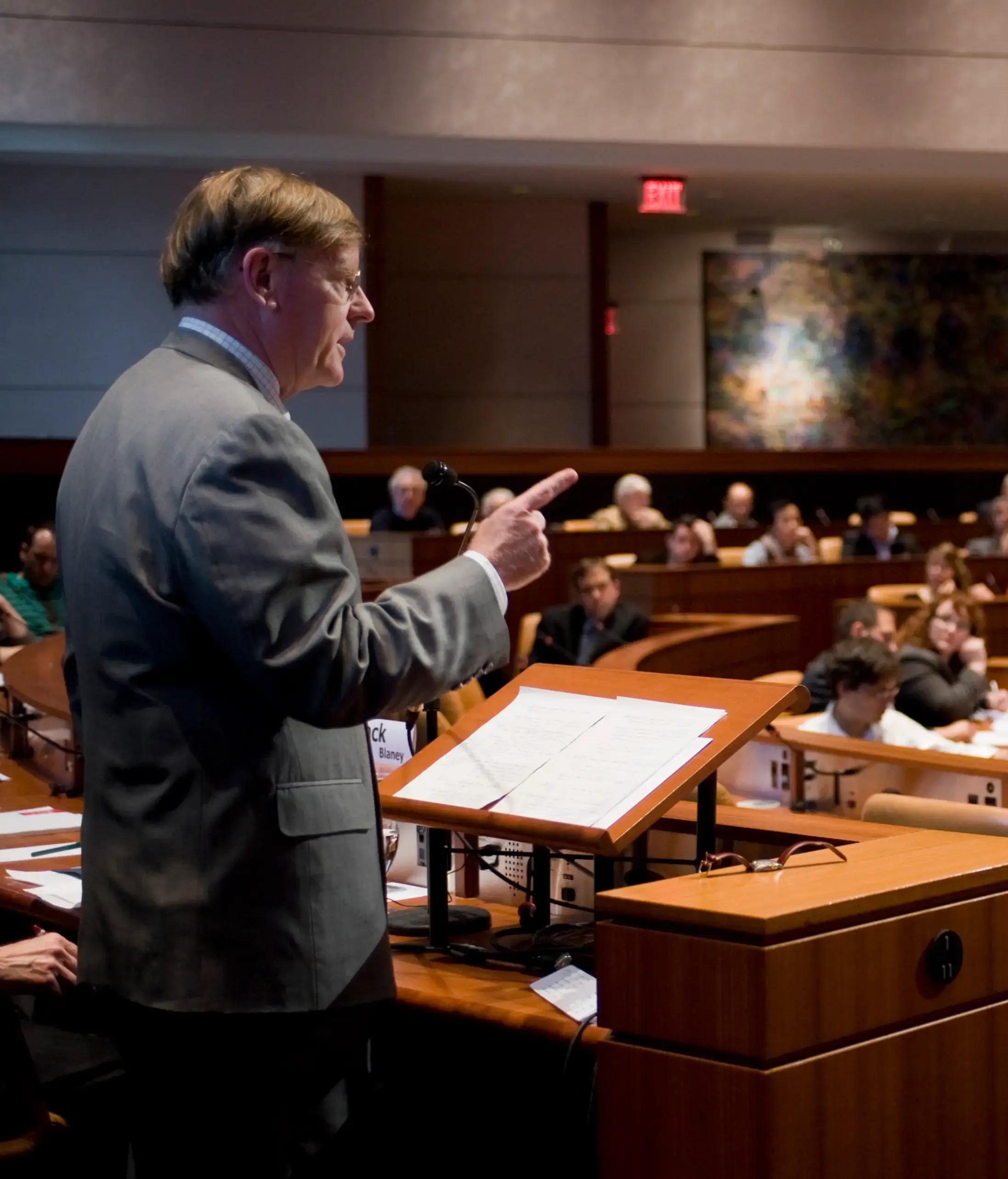 A person in a suit speaking at a podium in a large conference room with an audience seated in curved rows.