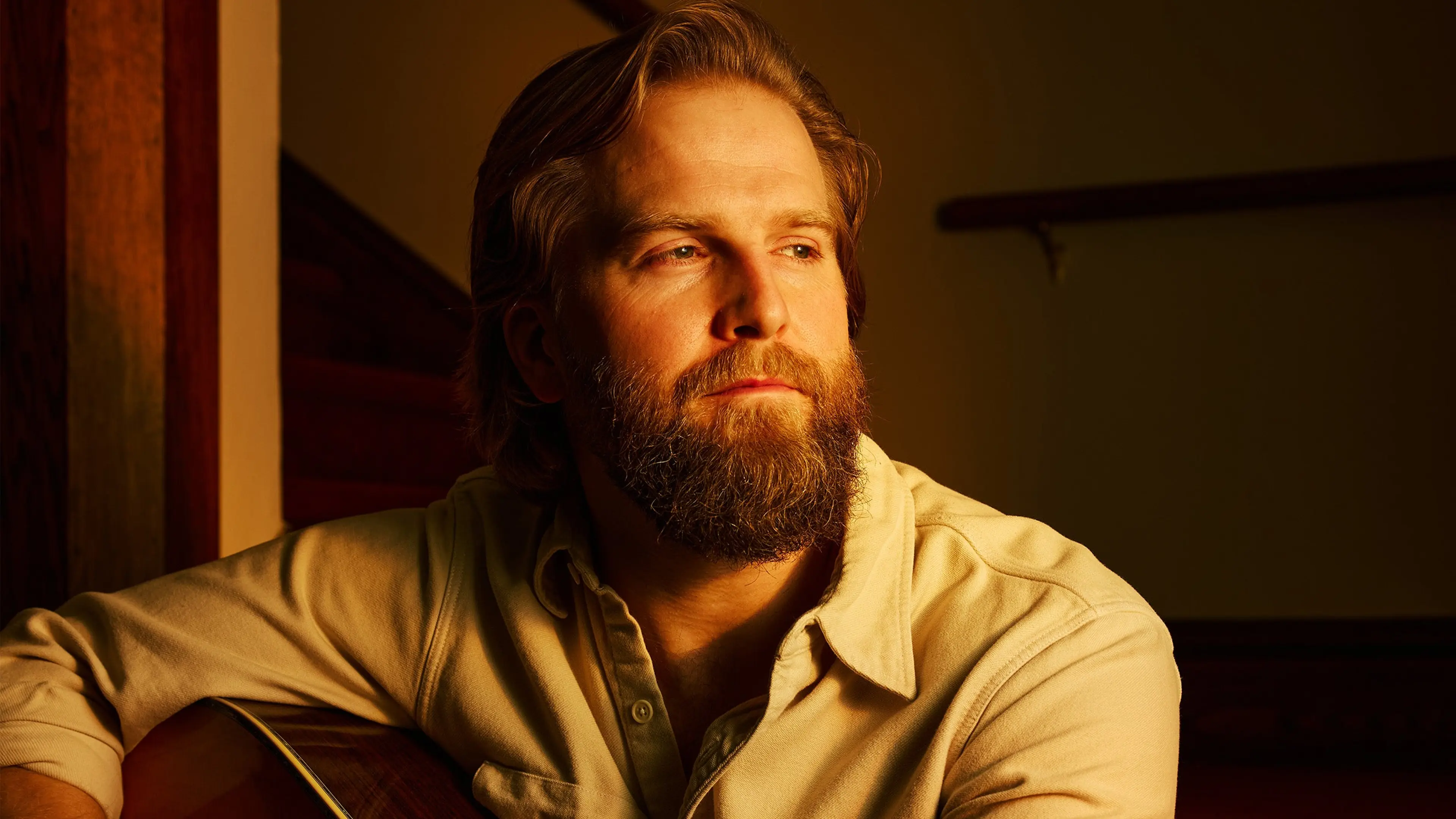 Person with beard and mustache wearing a beige shirt, sitting and holding a guitar, looking to the side in warm lighting.