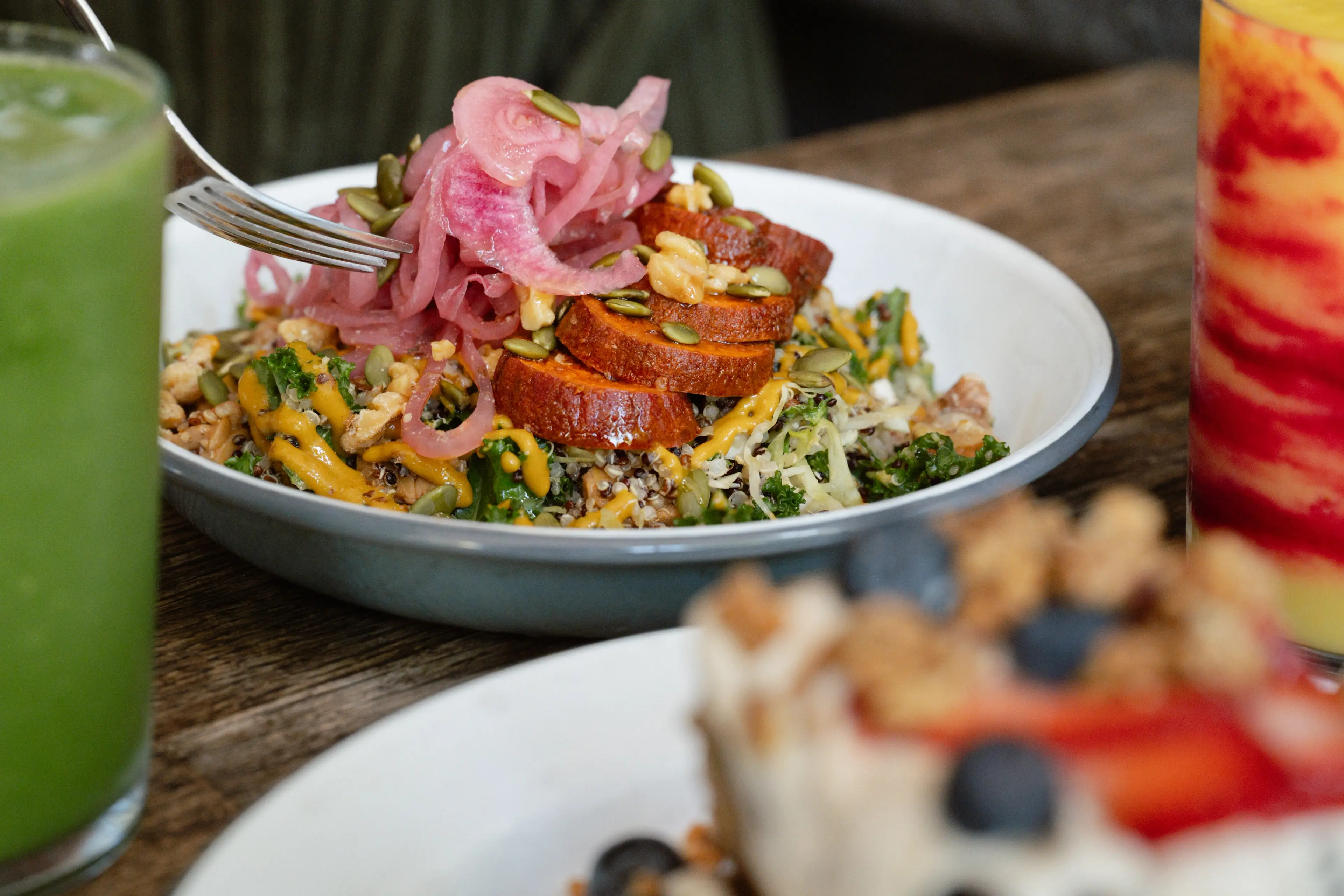 Bowl of salad with sliced sweet potatoes, pickled onions, pumpkin seeds, and dressing on a wooden table with green and orange drinks nearby.