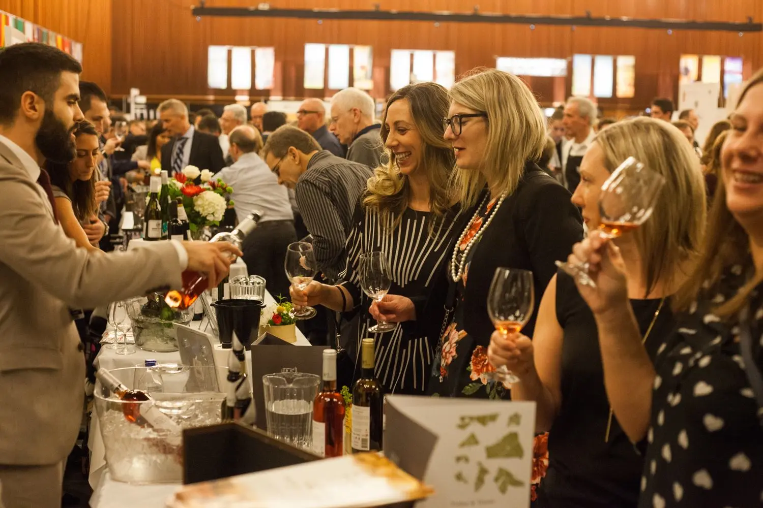 People at a wine tasting event holding glasses while a person pours wine from a bottle behind a table with wine bottles and flowers.