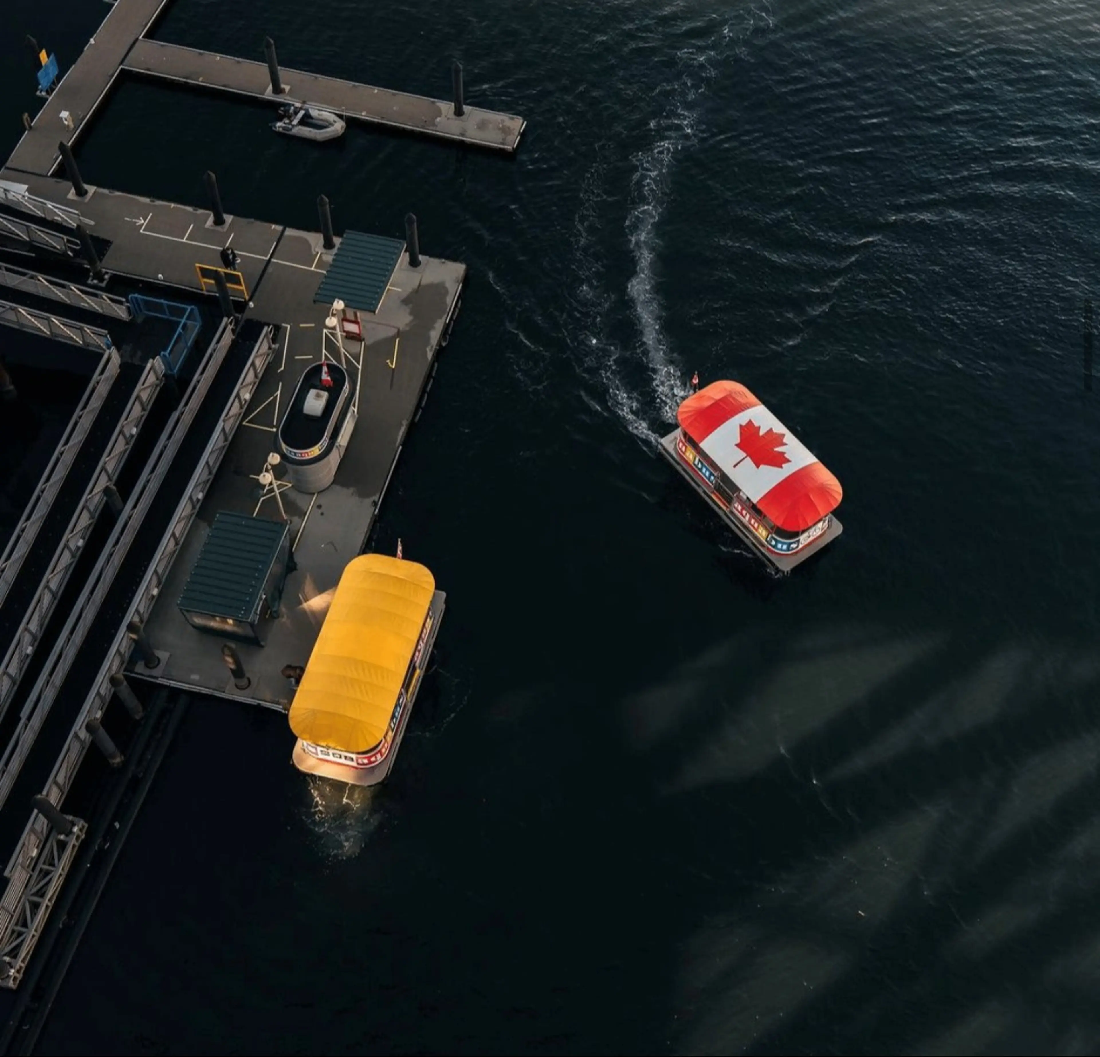 Aerial view of two small boats with colorful canopies, one with a Canadian flag, near a dock on dark water.