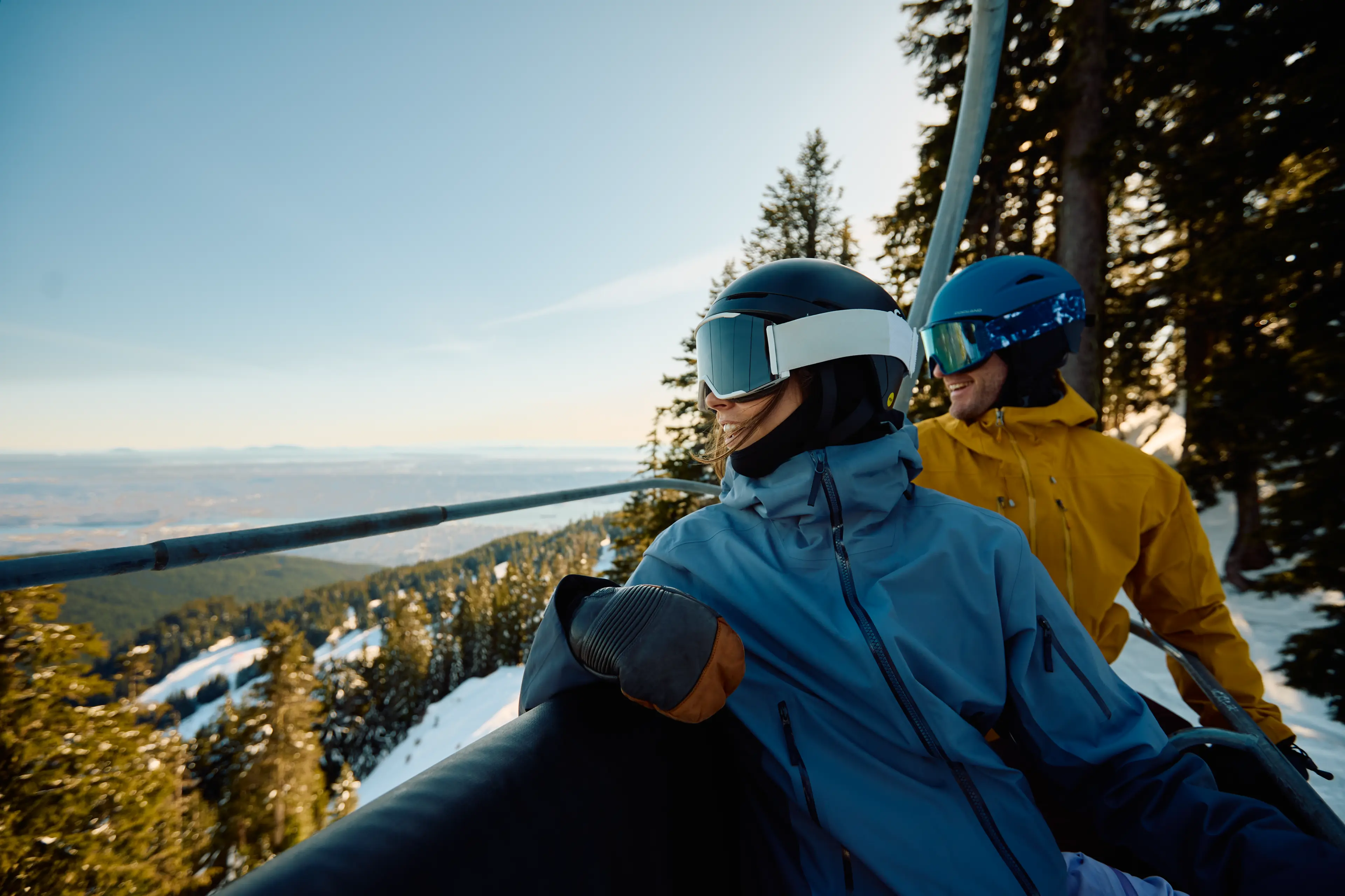 Two people in ski gear and helmets riding a ski lift over a snowy mountain with trees.