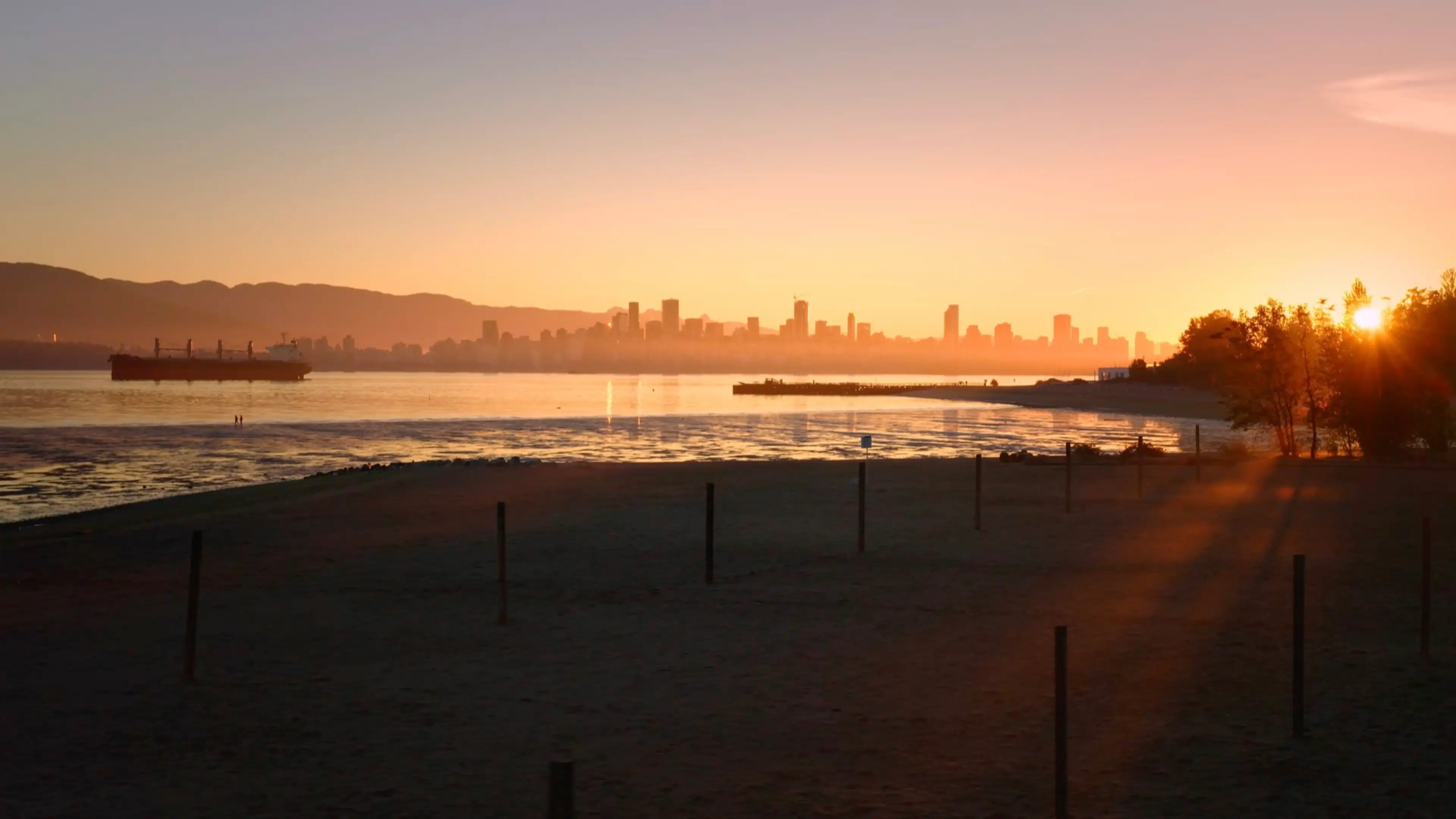 Sunset over a beach with silhouetted city skyline, a ship on the water, and sun rays through trees on the right
