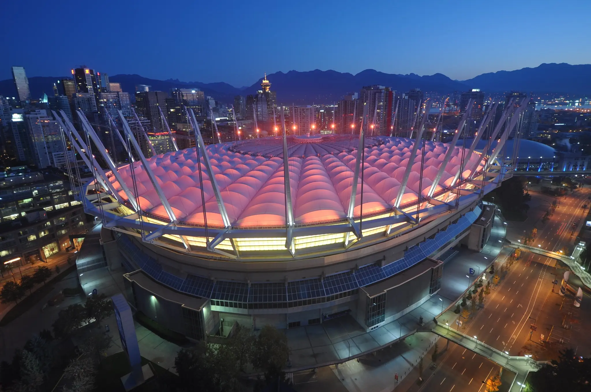 A glowing pink stadium with metal beams pointed upwards towers over the street.