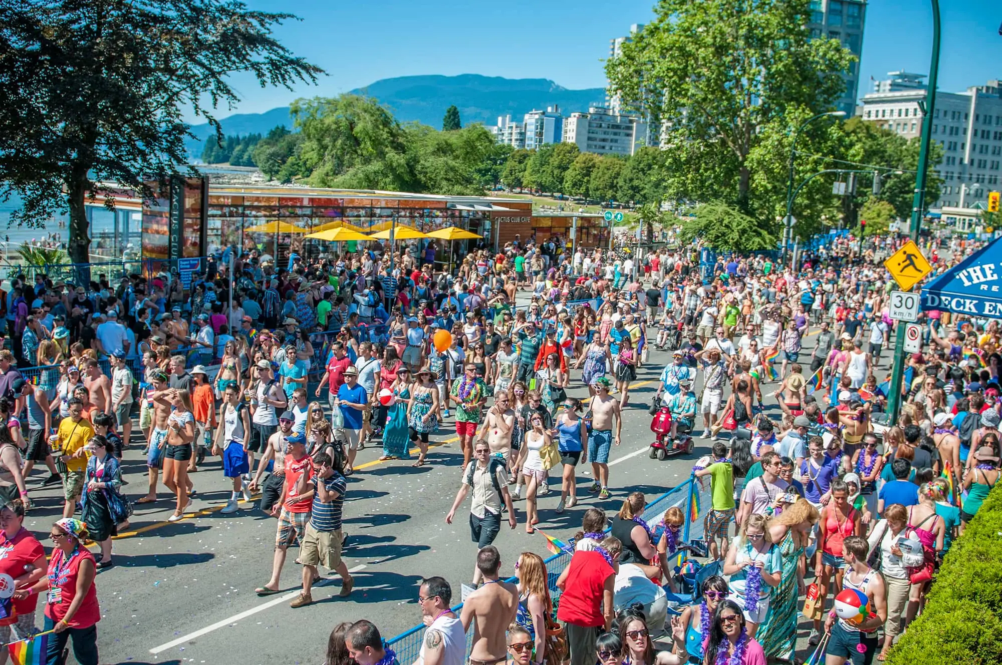 Large crowd of people walking and gathering outdoors on a sunny day near trees and buildings.