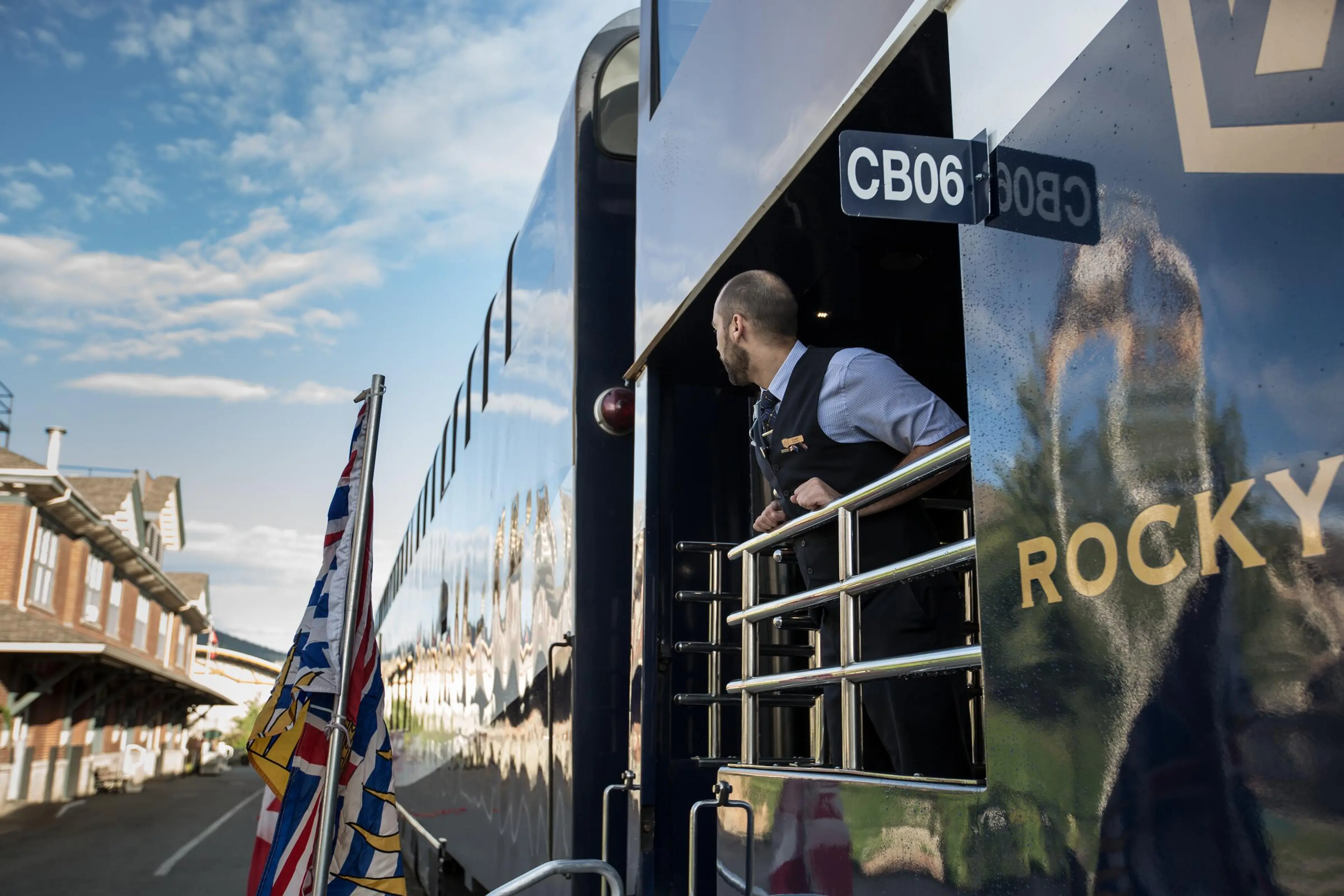 Person leaning out of a blue train car labeled CB06 and ROCKY, with flags and a station platform visible.