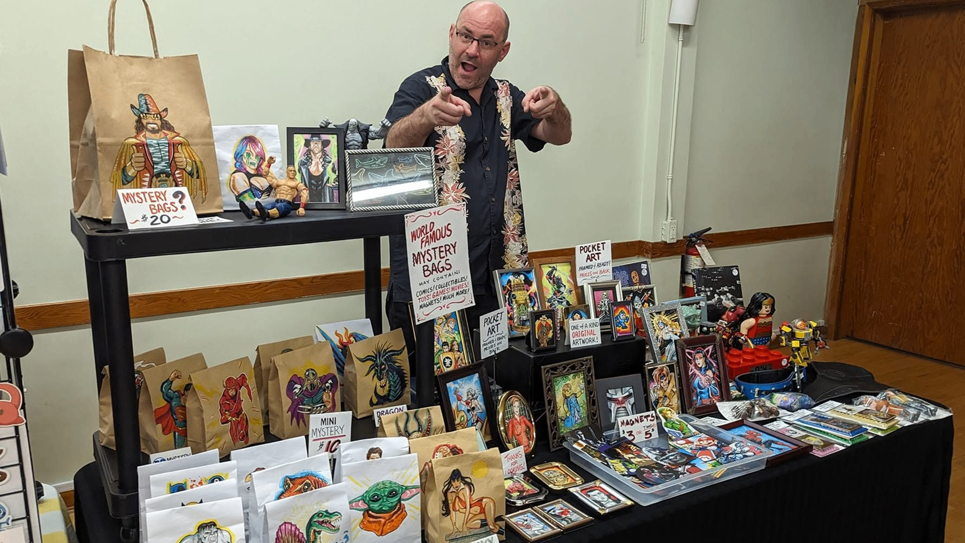 Person behind table with mystery bags, framed artwork, and collectibles displayed for sale at an indoor event.