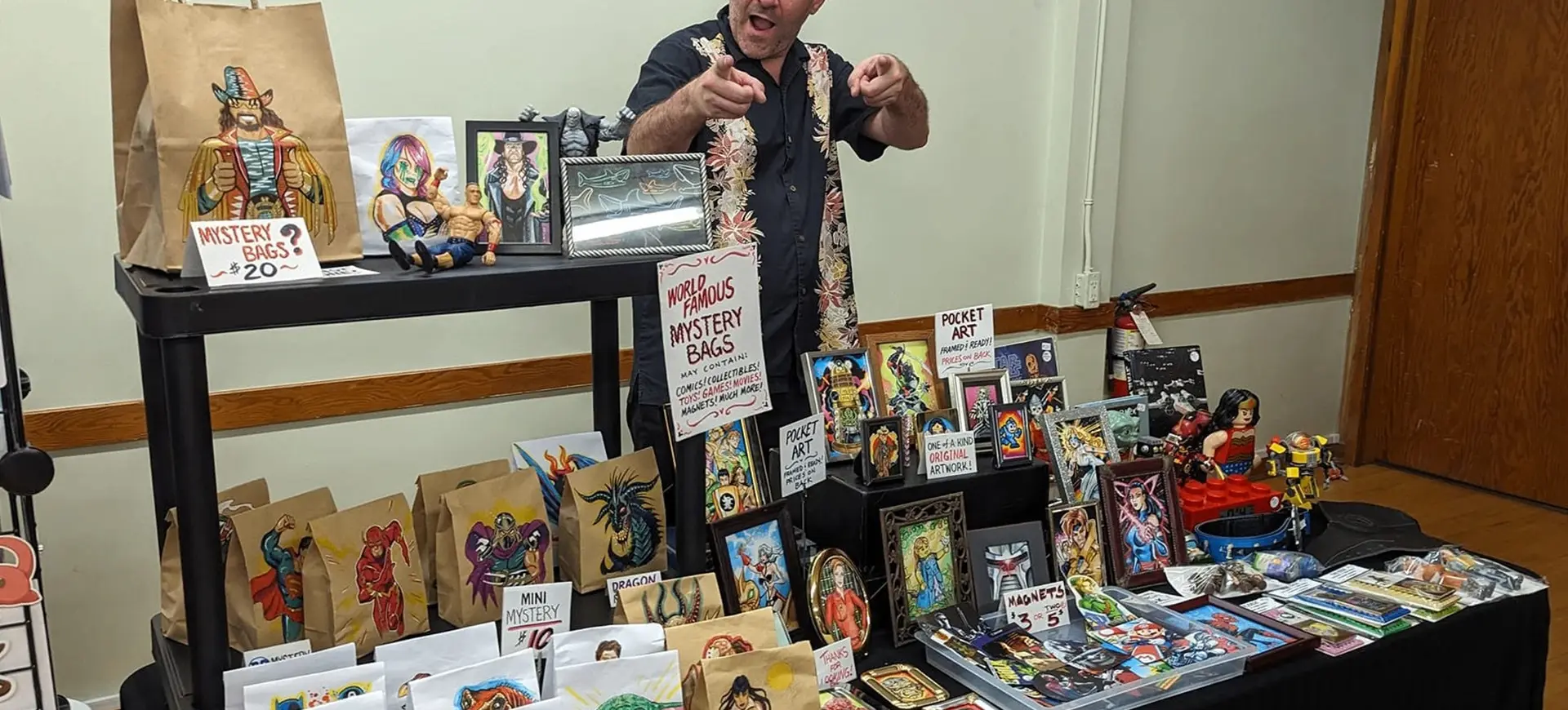 Person behind table with mystery bags, framed artwork, and collectibles displayed for sale at an indoor event.