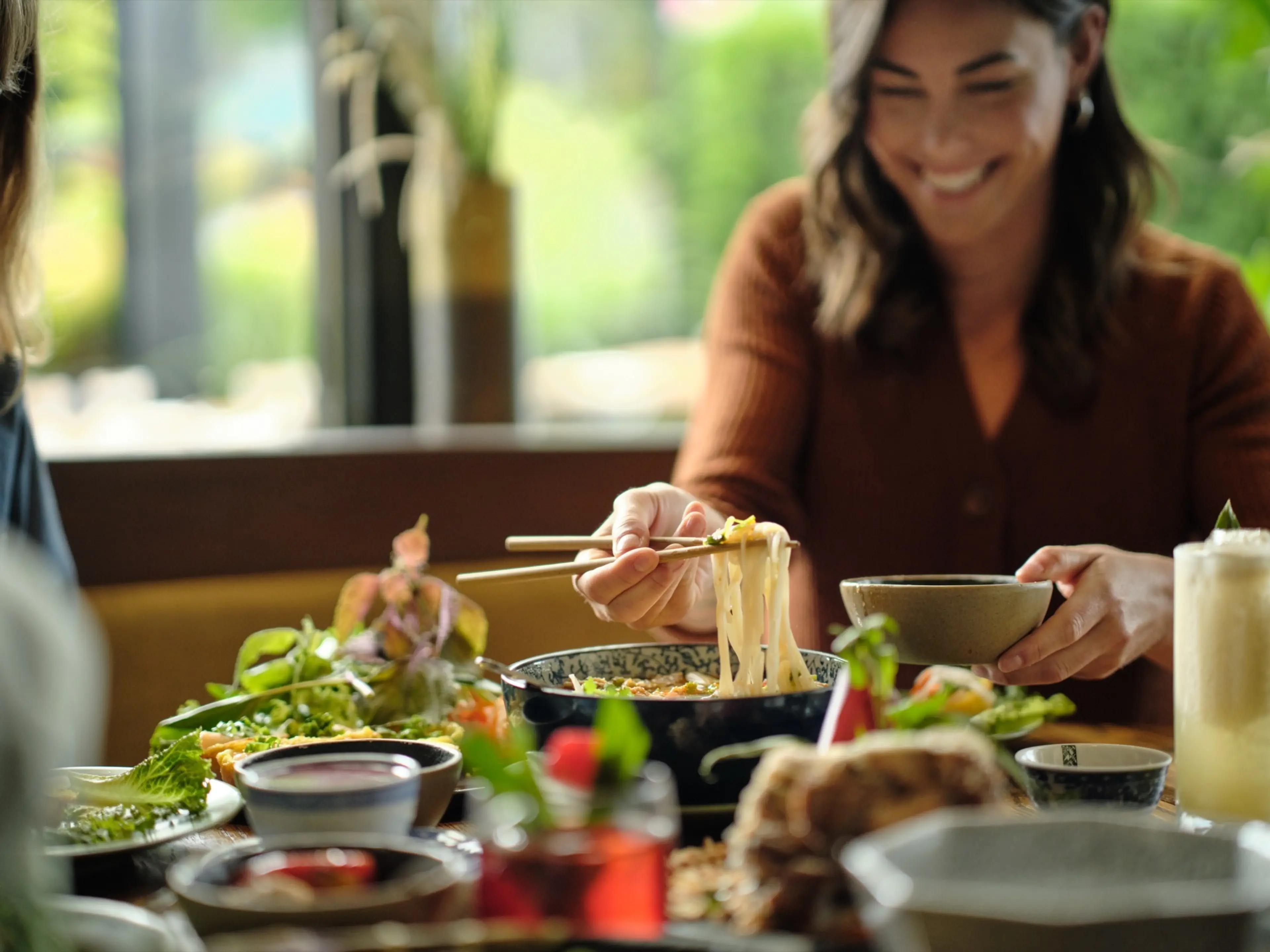 Smiling woman using chopsticks to pick up noodles from a bowl at a table with various dishes and drinks.