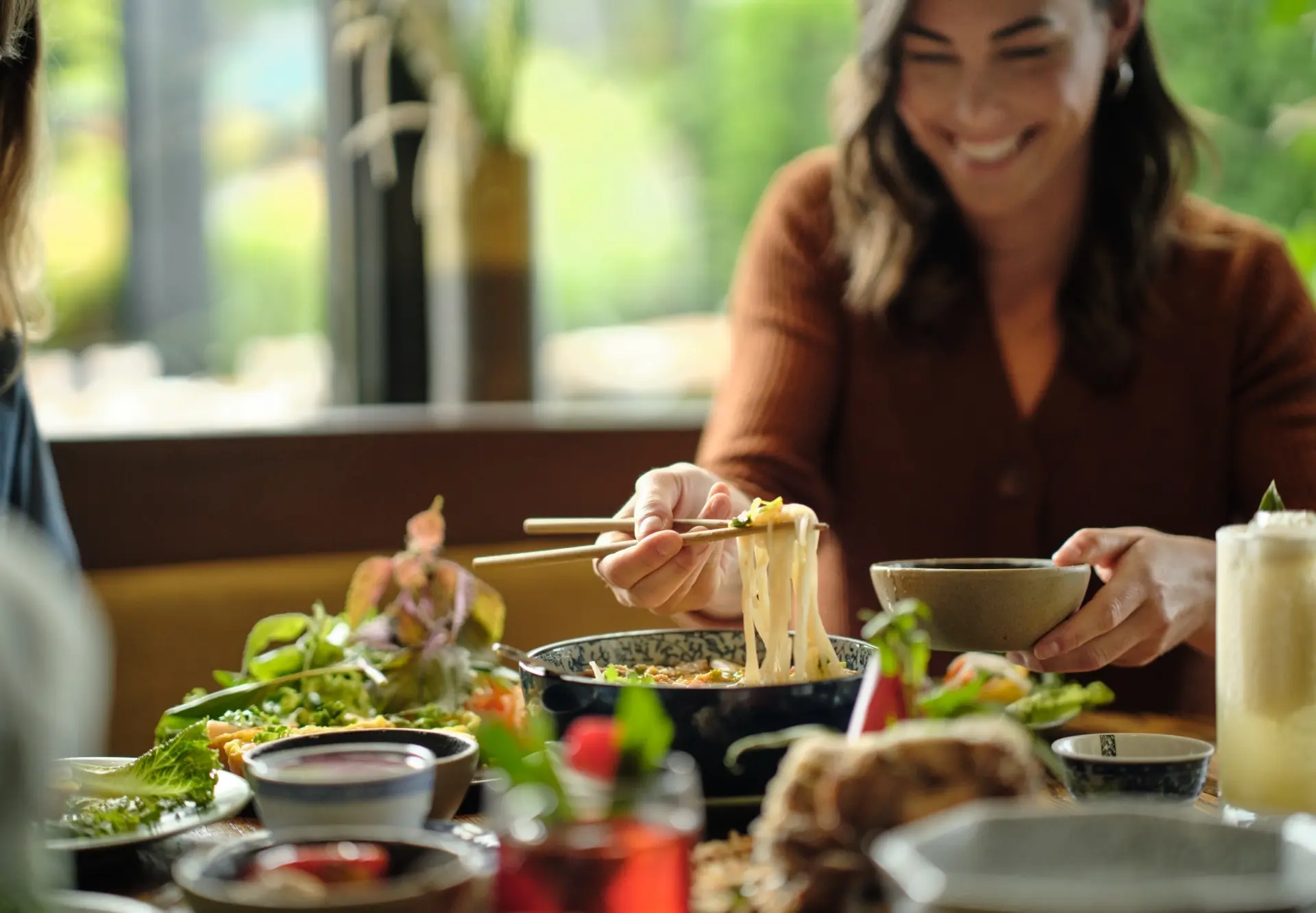 Person using chopsticks to lift noodles from a bowl at a table with various dishes and drinks.
