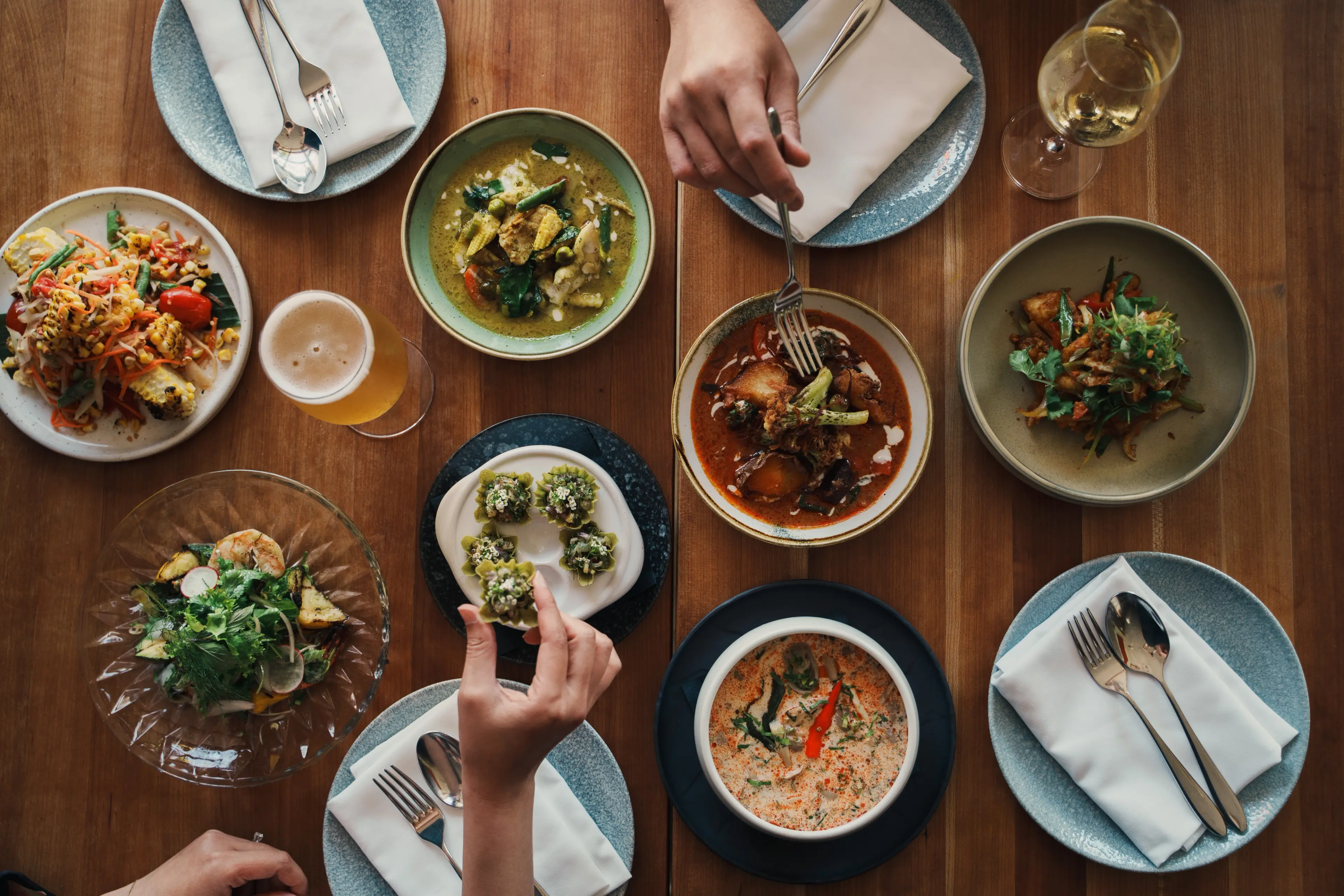 Overhead view of a wooden table with various dishes, two hands reaching for food, and drinks including a beer and a glass of white wine.