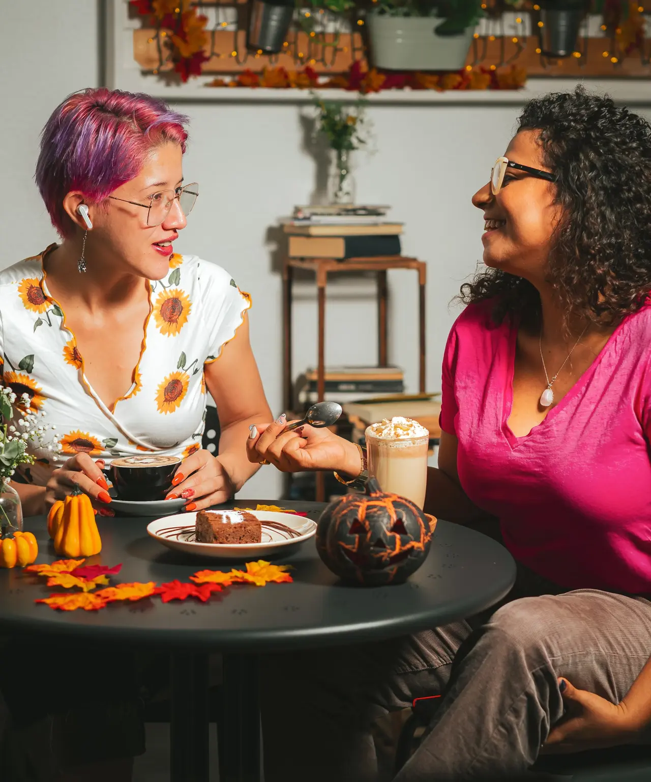 Two women sit at a table decorated with autumn leaves, pumpkins, and desserts, smiling and talking about a Vancouver itinerary. One has short pink hair and a sunflower dress; the other wears glasses and a pink shirt. Cozy, festive indoor setting.