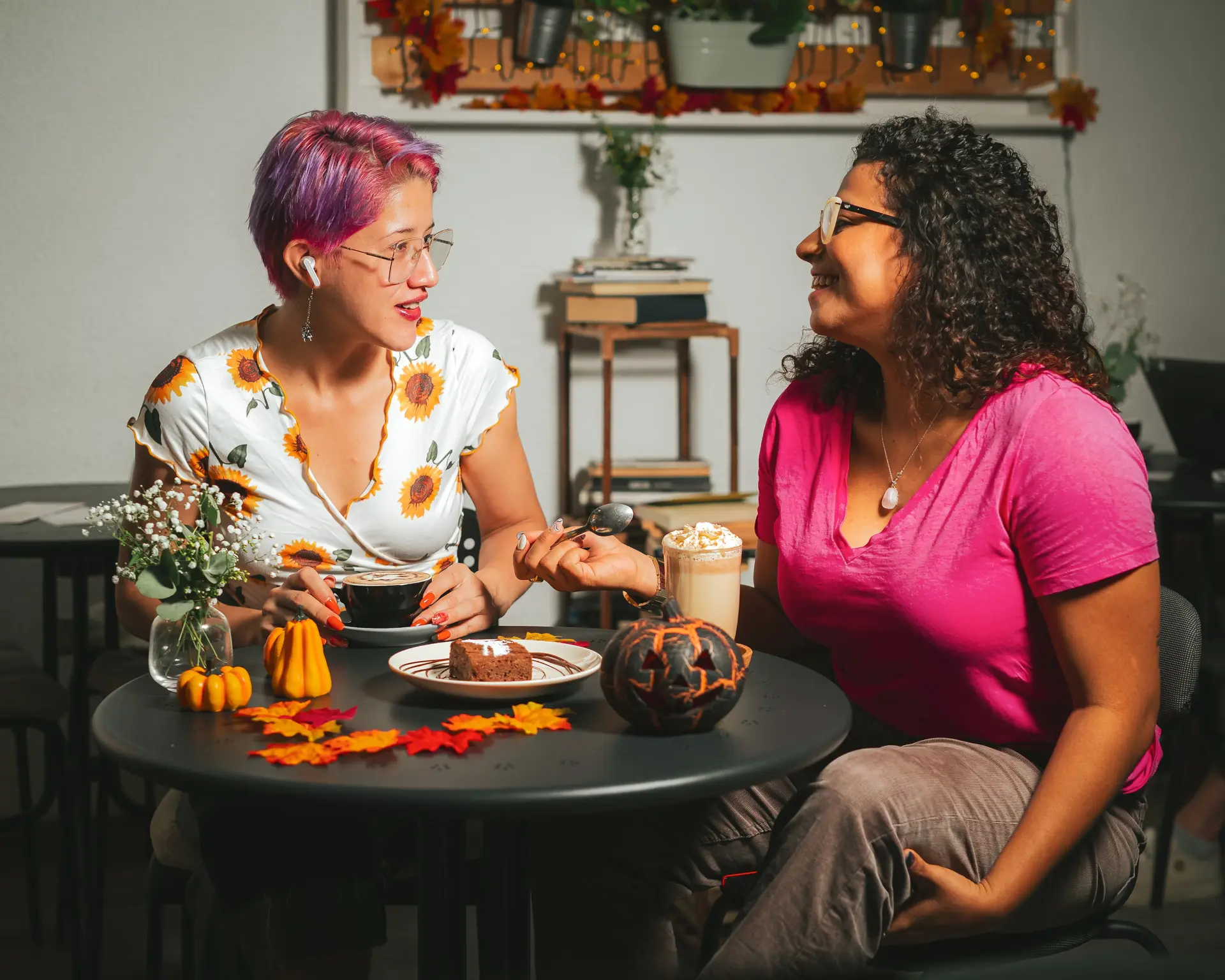 Two women sit at a table decorated with autumn leaves, pumpkins, and desserts, smiling and talking about a Vancouver itinerary. One has short pink hair and a sunflower dress; the other wears glasses and a pink shirt. Cozy, festive indoor setting.