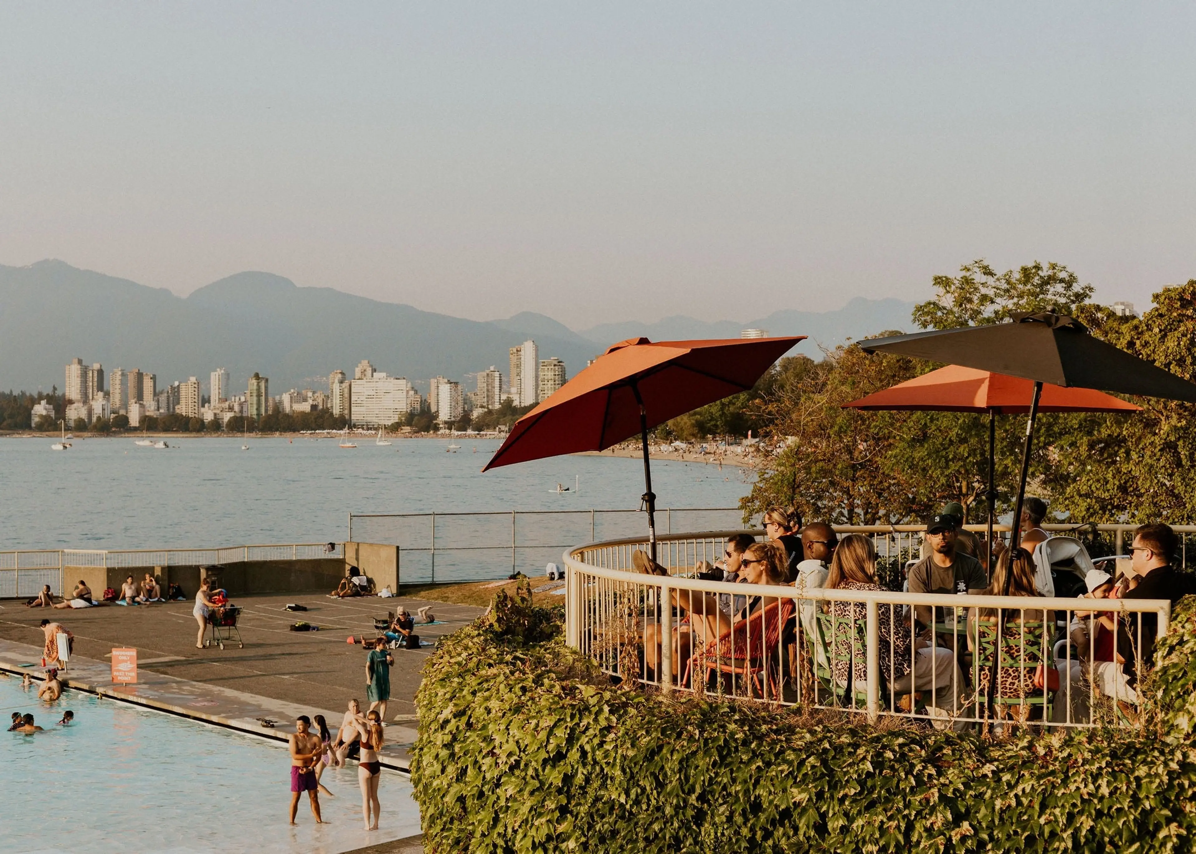 People sitting under umbrellas on a terrace covered with green vines, overlooking a pool and a city skyline with mountains in the background.